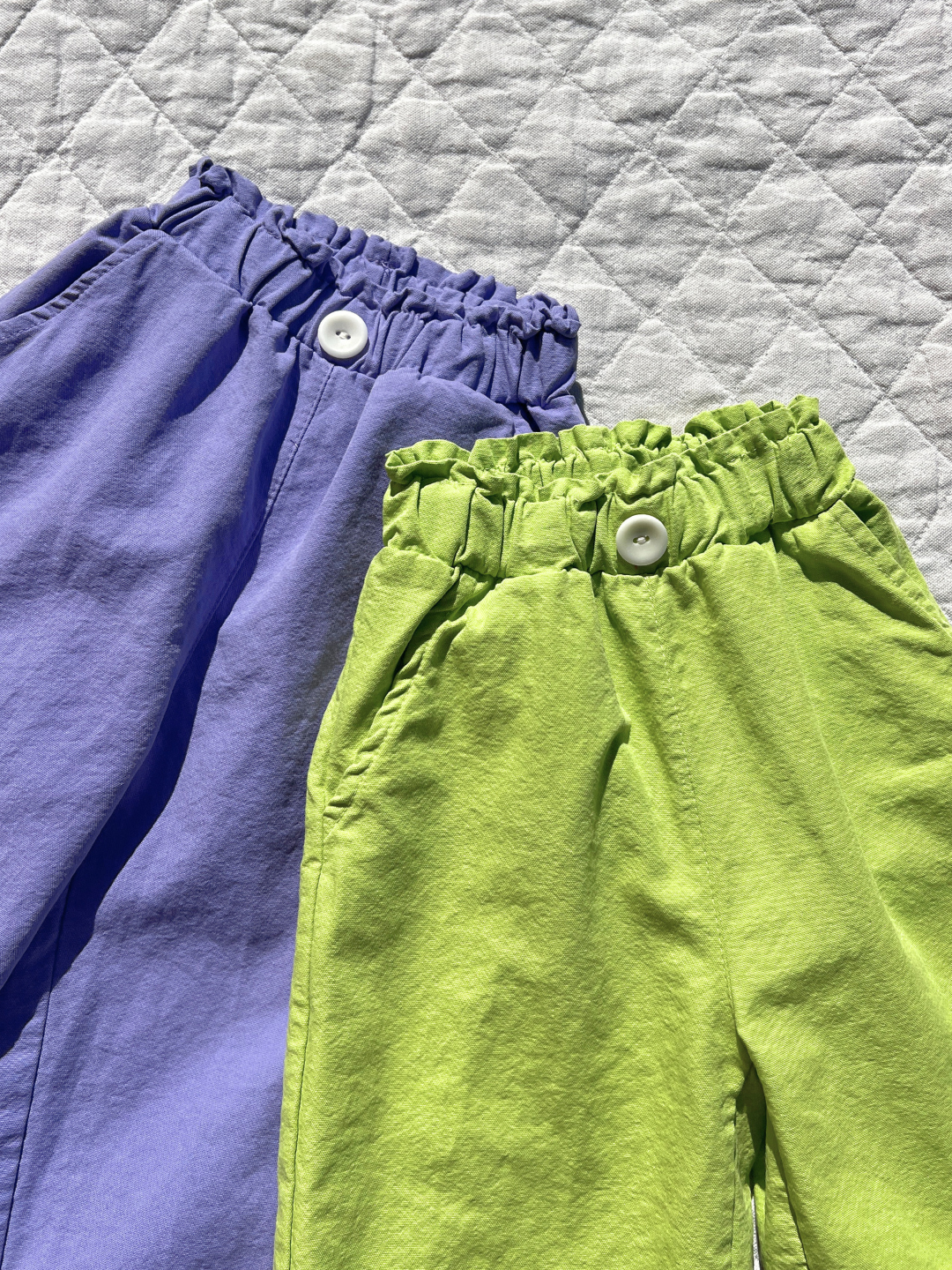 Green | The JELLY PANTS, purple and green relaxed pull-on pants from a Korean kids brand, are crafted from woven cotton with elastic waistbands and white buttons, displayed on a quilted white surface.
