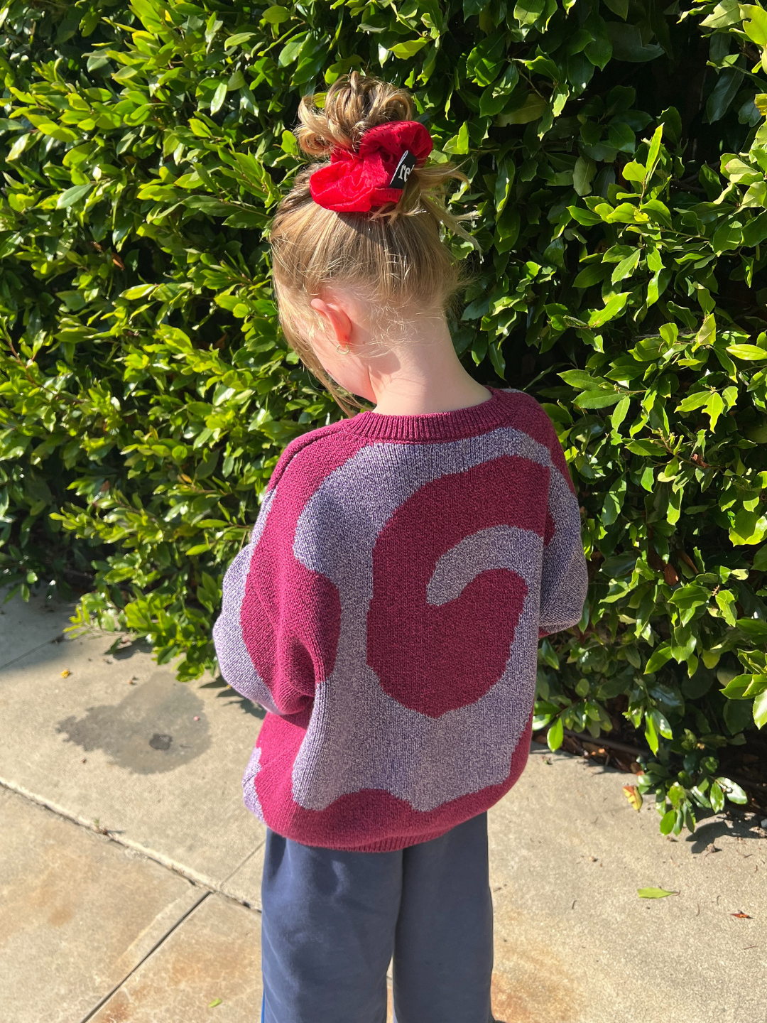 Pink/Purple | A child wearing the SWIRL SWEATER—with a purple and pink swirl pattern—stands on a sidewalk in front of green bushes, facing away from the camera and sporting a red hair scrunchie.