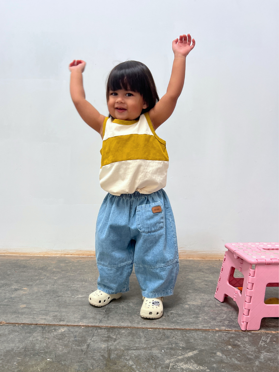 Cream/Gold | A young child in a relaxed fit TRACK TANK and blue jeans stands indoors with arms raised, next to a small pink plastic step stool.