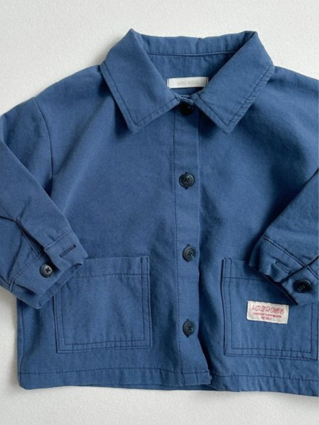 Blue | The MINI WORKWEAR JACKET is a blue kids’ button-up with a collar, two front pockets, and a small label on the lower right pocket, shown on a light background.