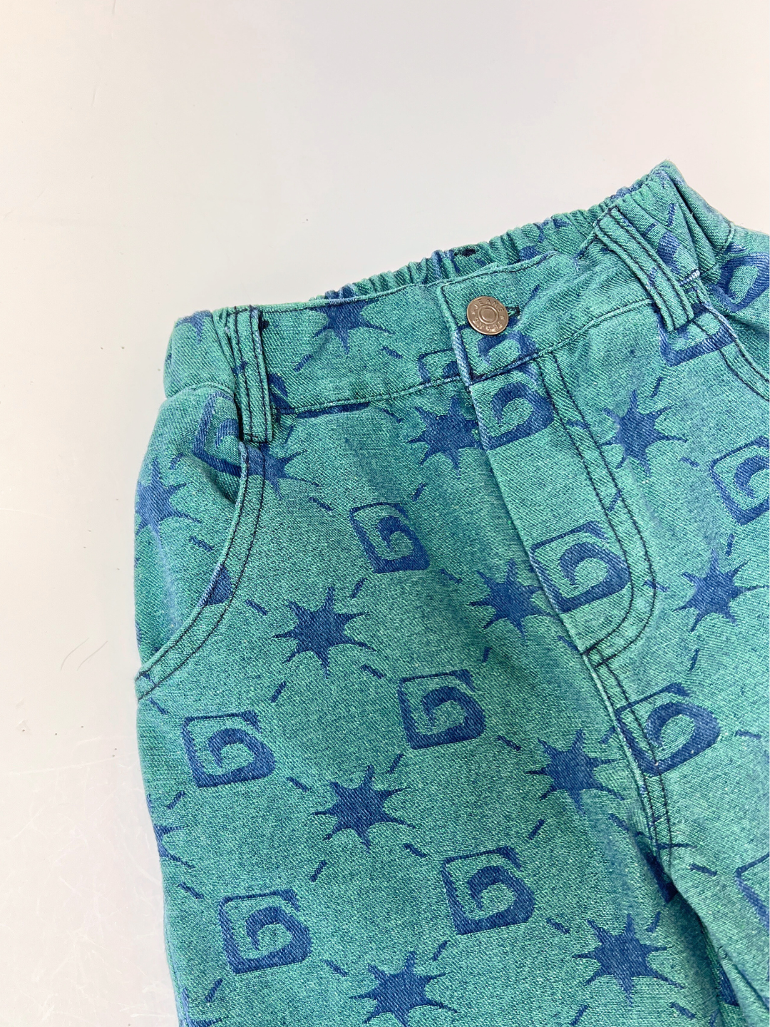 A close-up of TRELLIS JEANS in green, relaxed fit, from the Milk Teeth collection, featuring blue abstract starburst and square patterns, front pockets, belt loops, and a button closure.