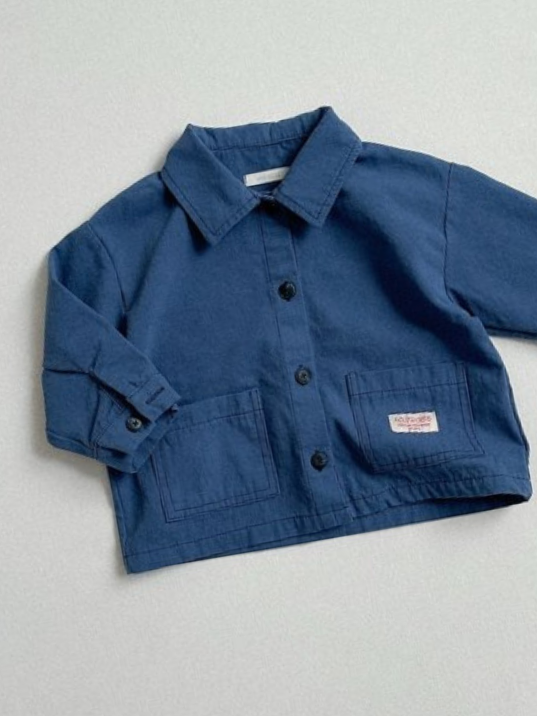 Blue | The MINI WORKWEAR JACKET is a blue, long-sleeve button-up with two front pockets and a small label on one pocket, shown laid flat on a light background.