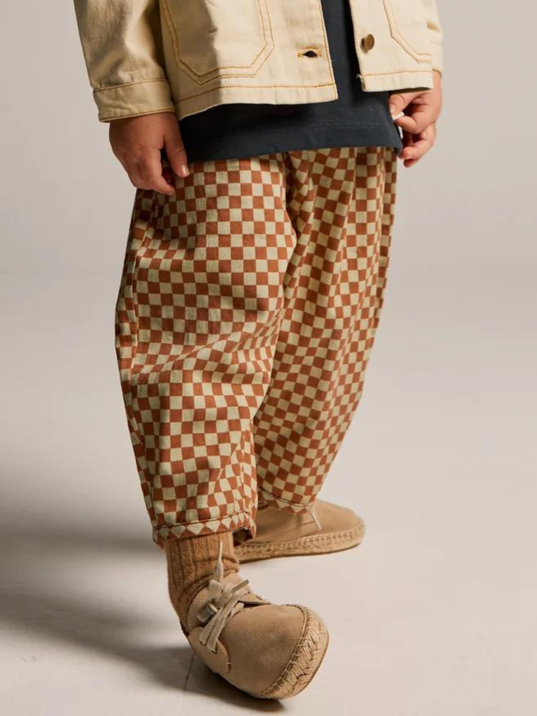 A child stands wearing BRICK CHECK TROUSER pants made from organic cotton twill, beige moccasin-style shoes, beige socks, a black shirt, and a beige jacket. The background is a plain light grey.