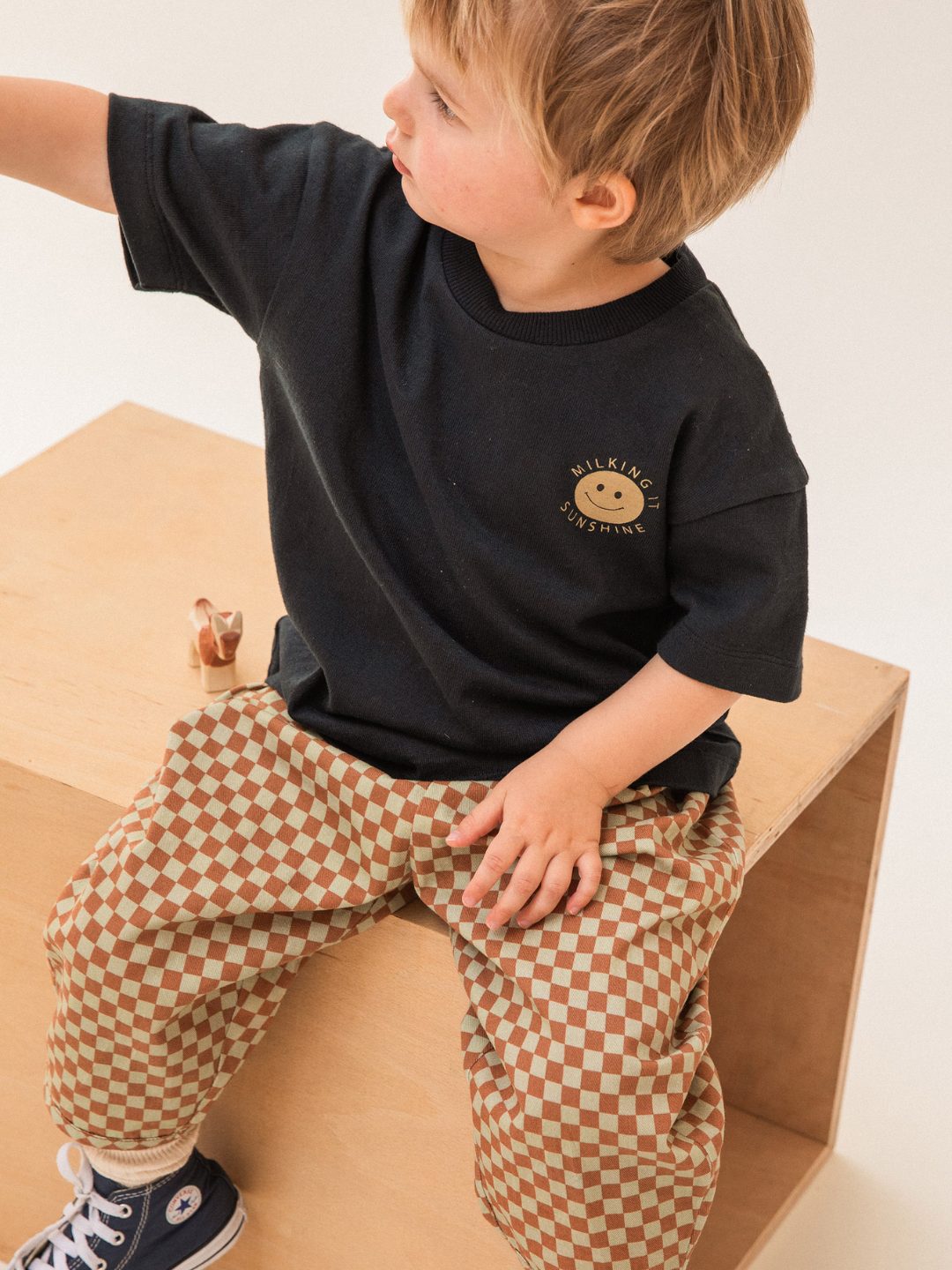 A young child with light hair sits on a wooden block. They are wearing a black t-shirt with a smiley face and the text "Walking Sunshine," BRICK CHECK TROUSER made from organic cotton twill, and dark sneakers. The child looks off to the side, reaching out with one arm.