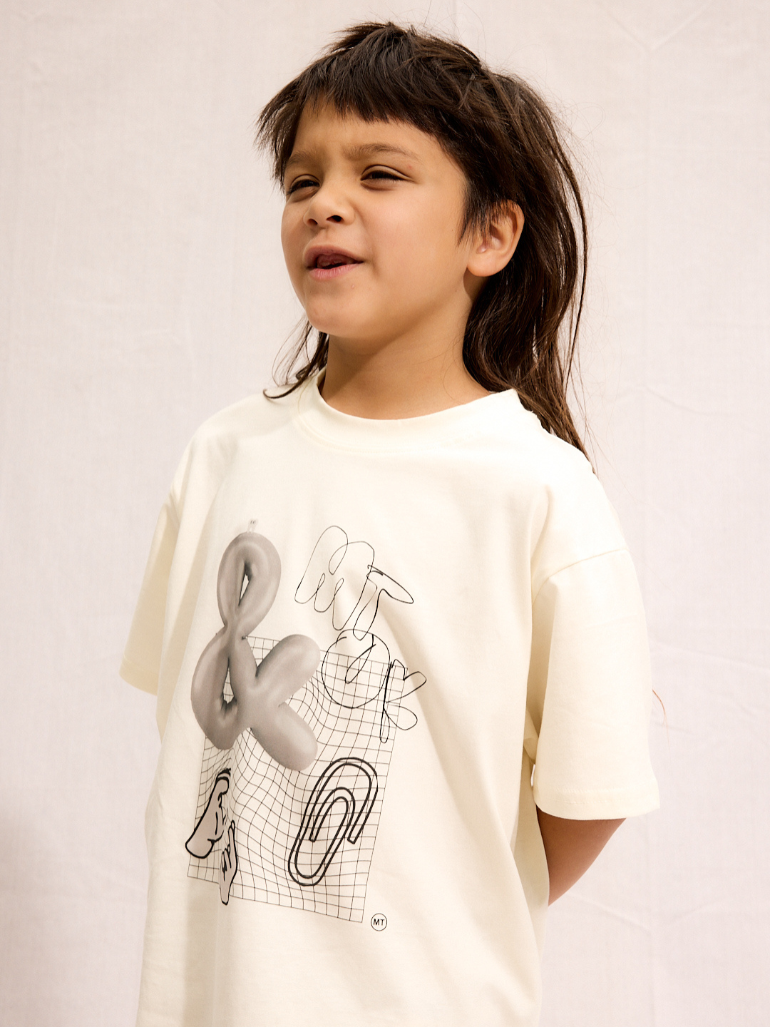 A child with long brown hair stands against a plain background, wearing the CLIP ART TEE—an oversized, silky-soft cream cotton shirt featuring abstract black and gray graphics.