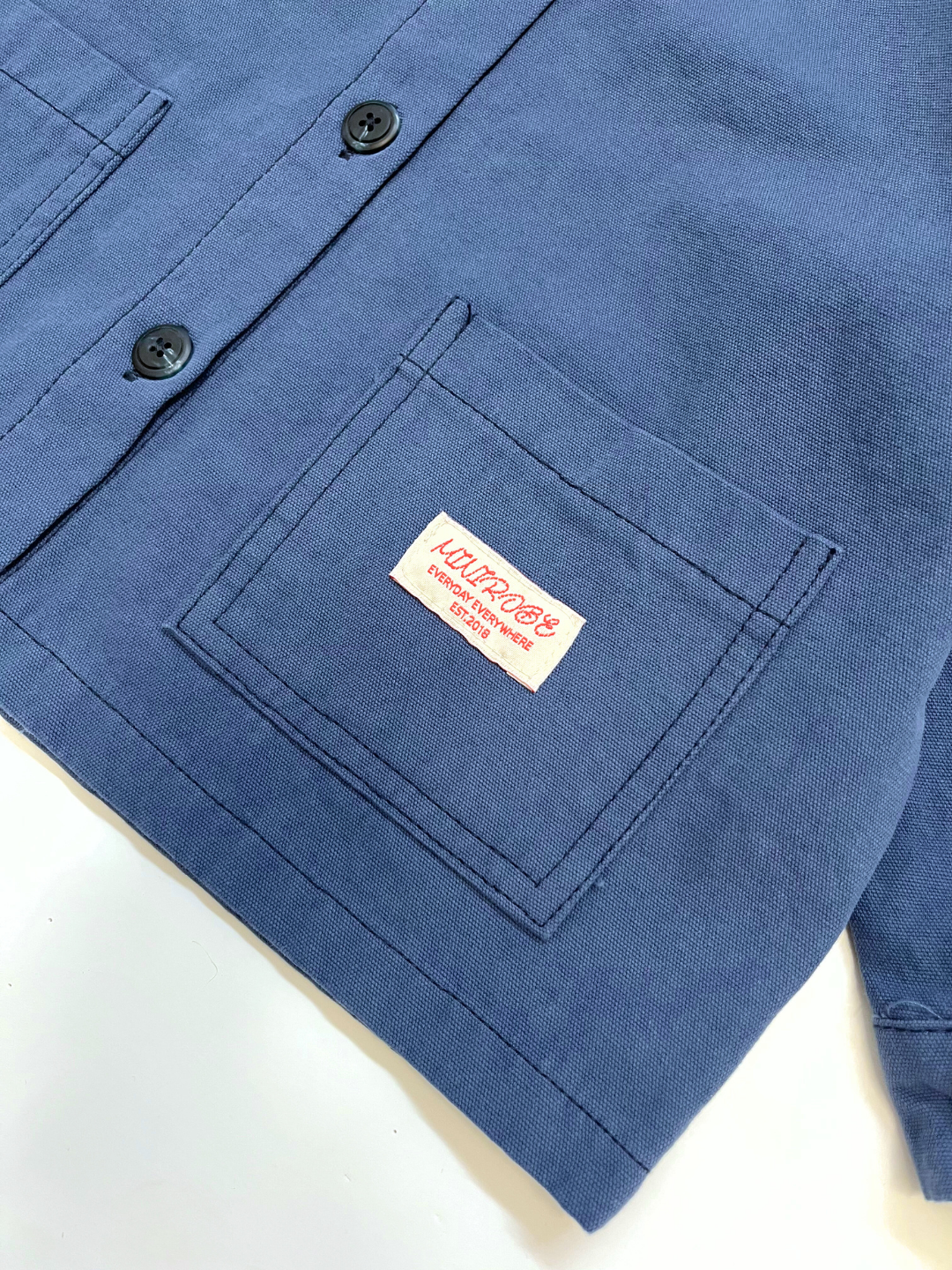 Blue | Close-up of the MINI WORKWEAR JACKET for kids in blue, showing a front pocket with a white and red tag labeled “Kurtrobe, Standard and Strong Goods.”.