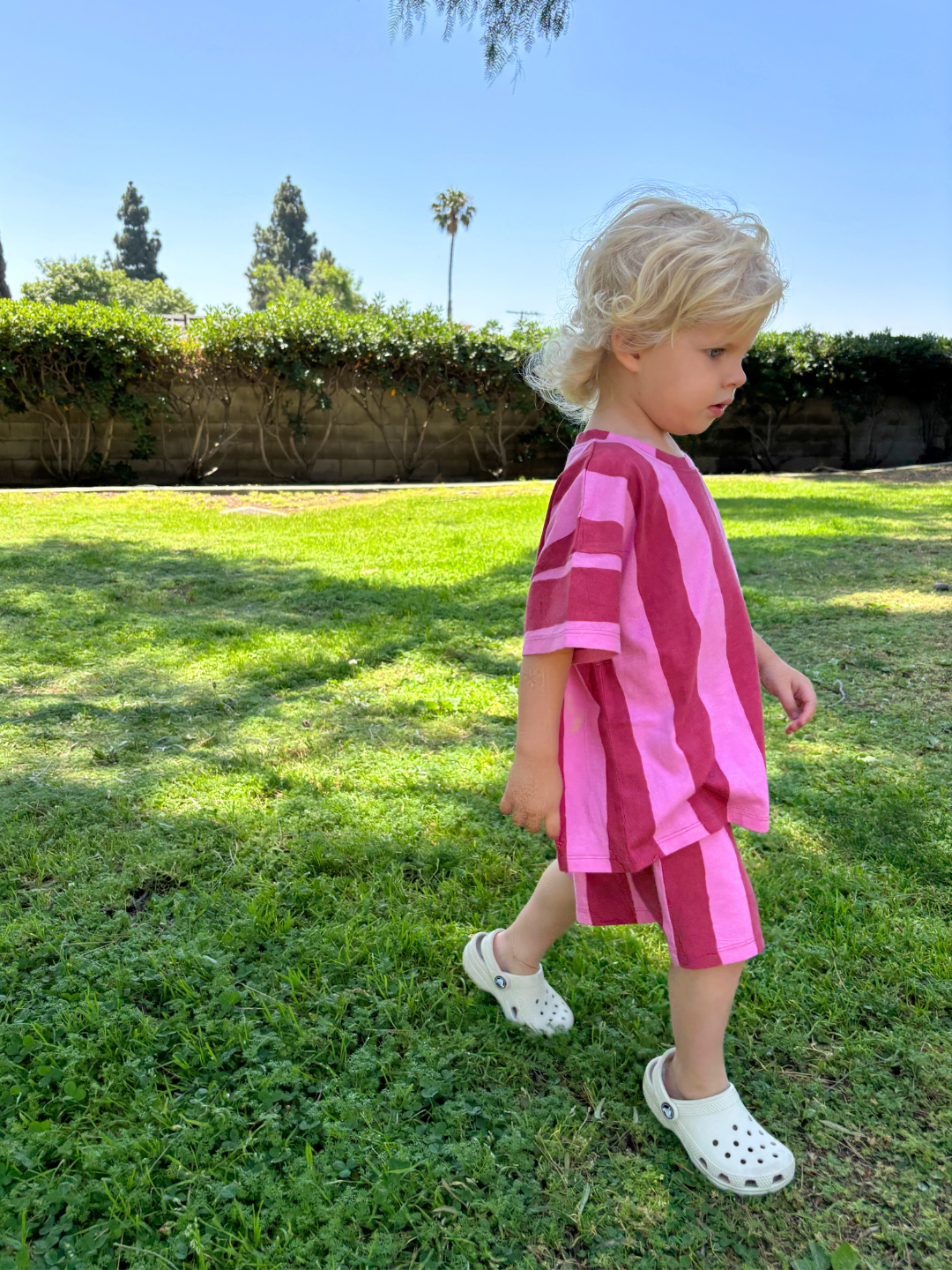 A young child walks on grass, wearing the UMBRELLA STRIPE SHORT SET in pink and red vertical stripes, made from soft organic cotton, paired with beige crocs, and blue sky behind.