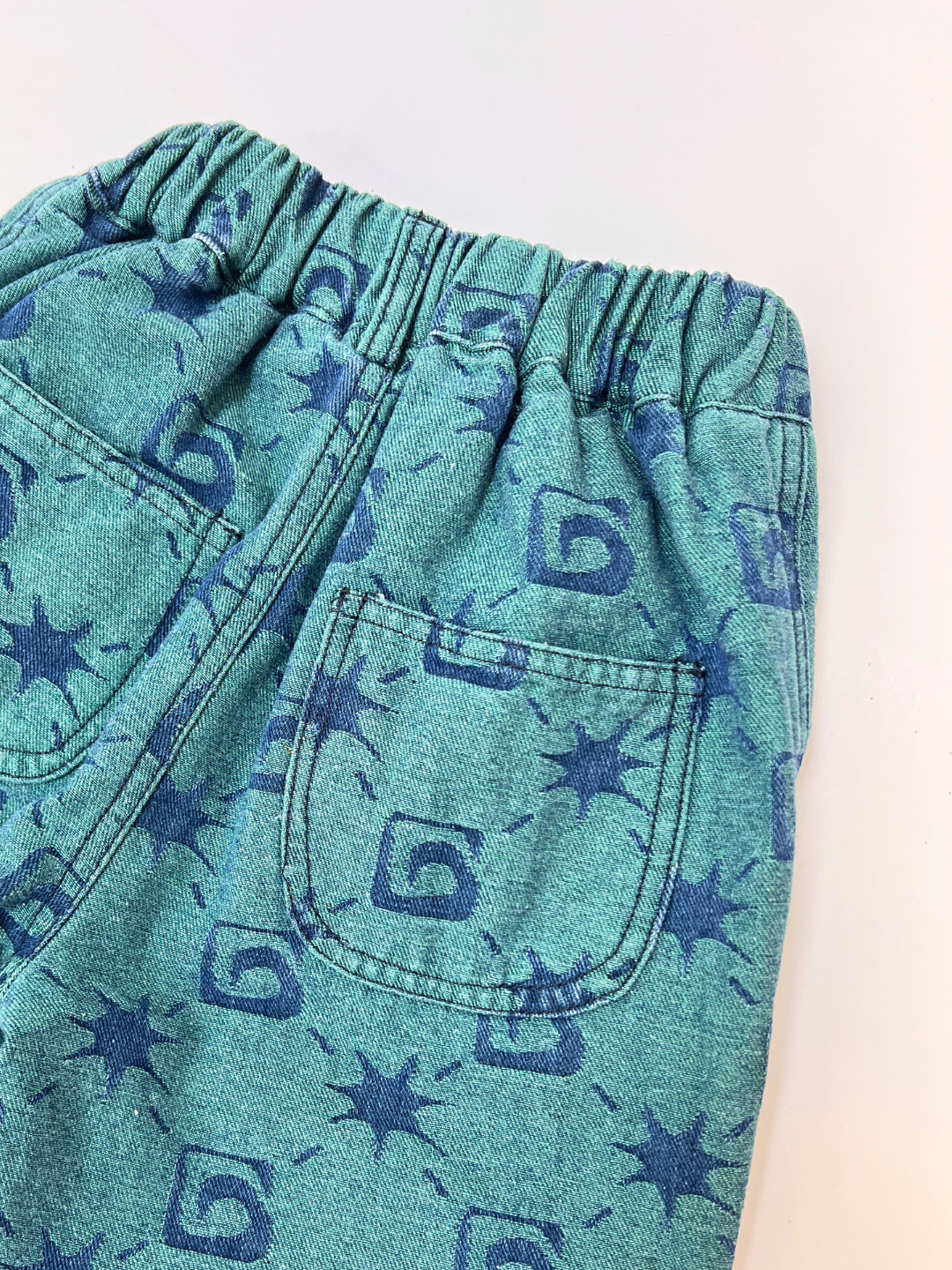 Close-up of TRELLIS JEANS: teal shorts with a single back pocket, elastic waistband, and a blue abstract pattern with spirals and star shapes.