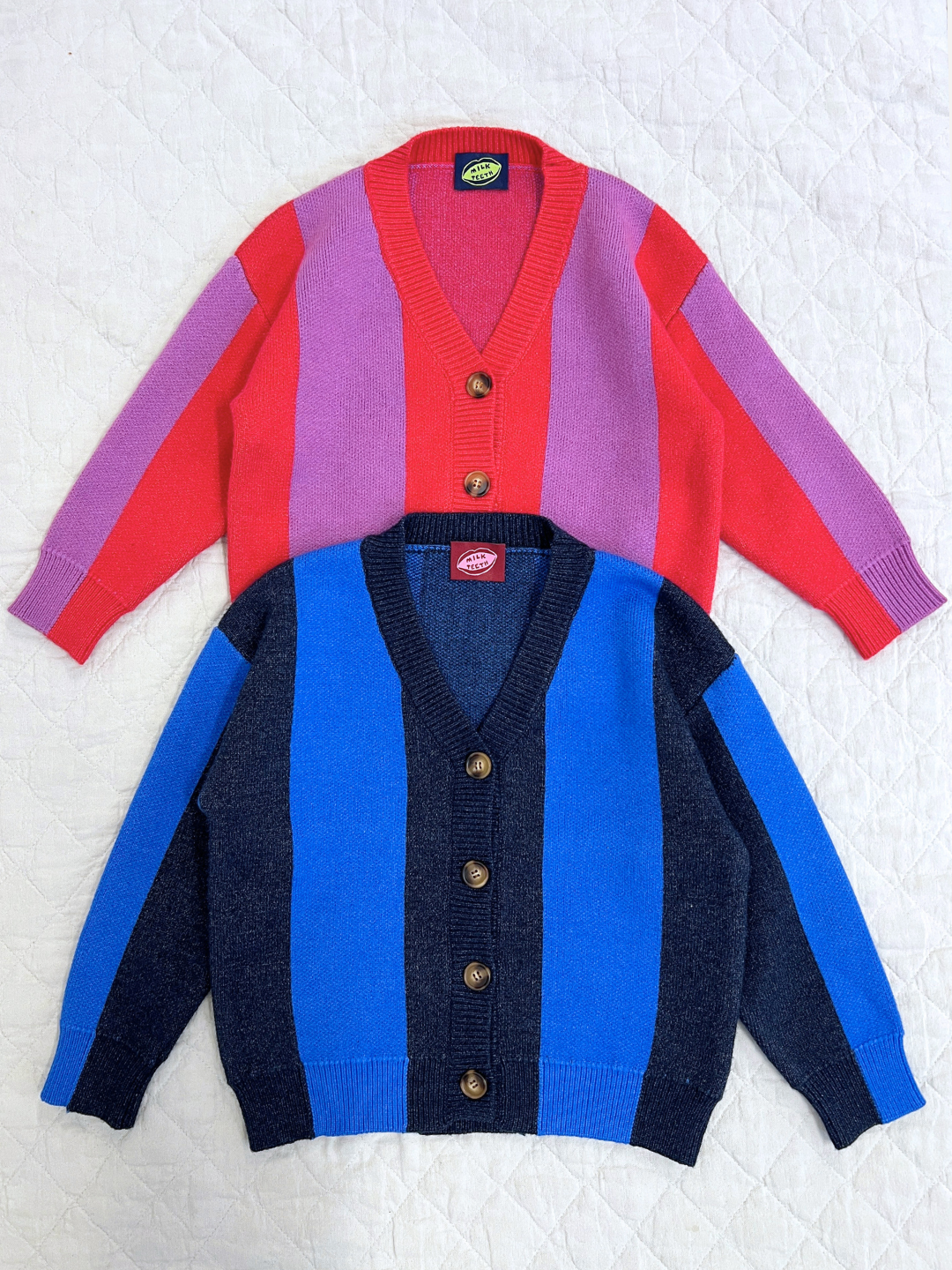 Red | Two STRIPE CARDIGANS with wide vertical stripes are laid flat on a quilted white surface: one is pink and red, the other is blue and navy.