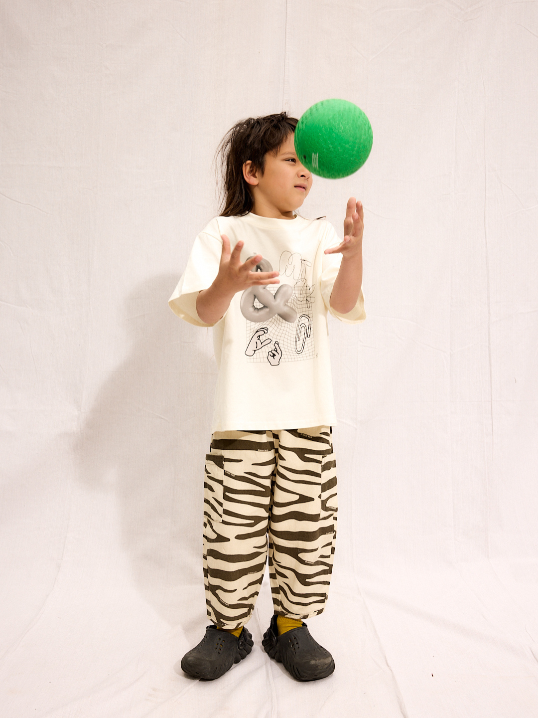 A young child stands on a white backdrop, tossing a green ball while wearing the CLIP ART TEE with a graphic print, zebra-striped pants, and black shoes.