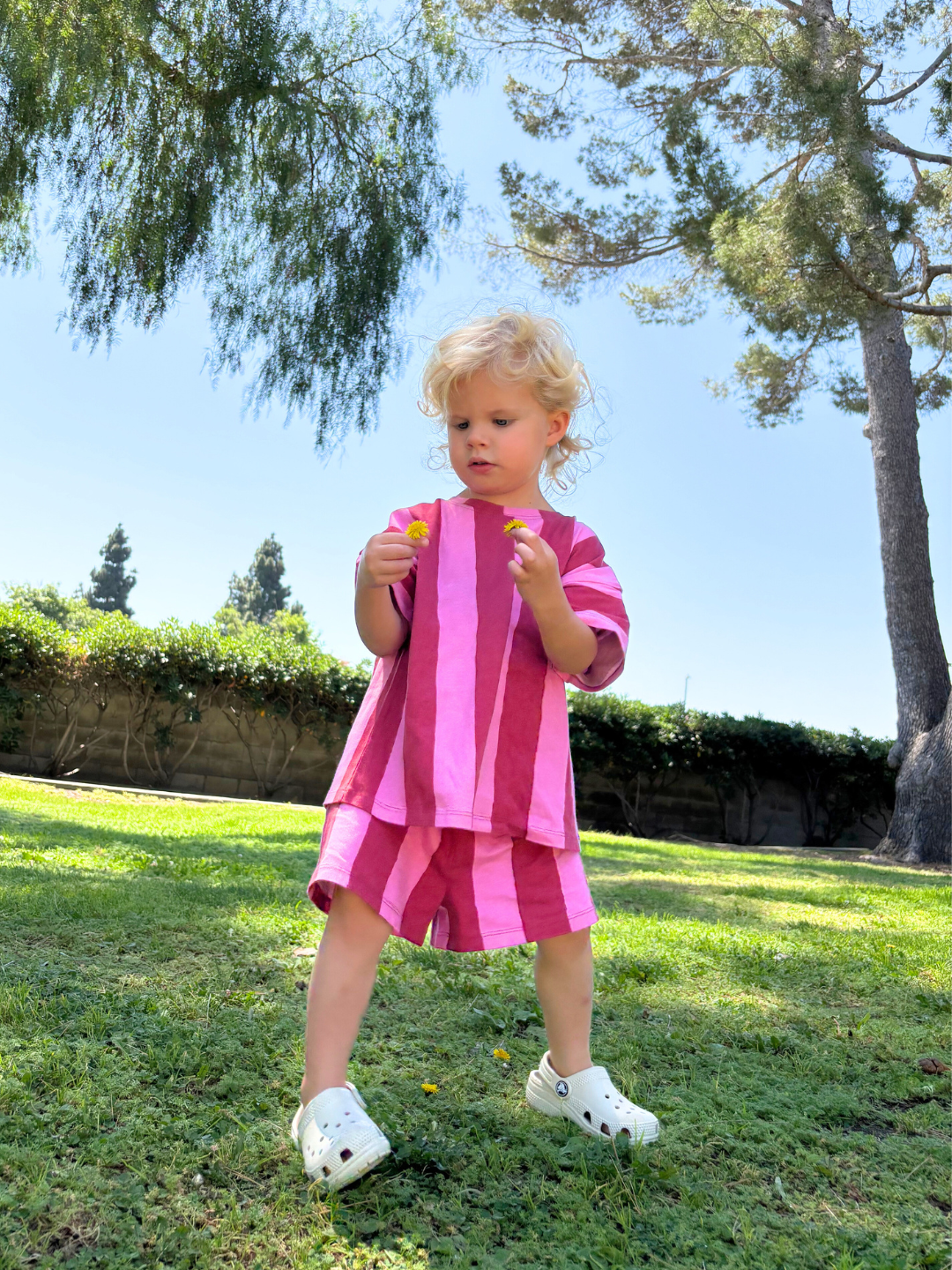 A young child walks on grass, wearing the UMBRELLA STRIPE SHORT SET in pink and red vertical stripes, made from soft organic cotton, paired with beige crocs, and blue sky behind. She has blonde hair and holds two flowers.