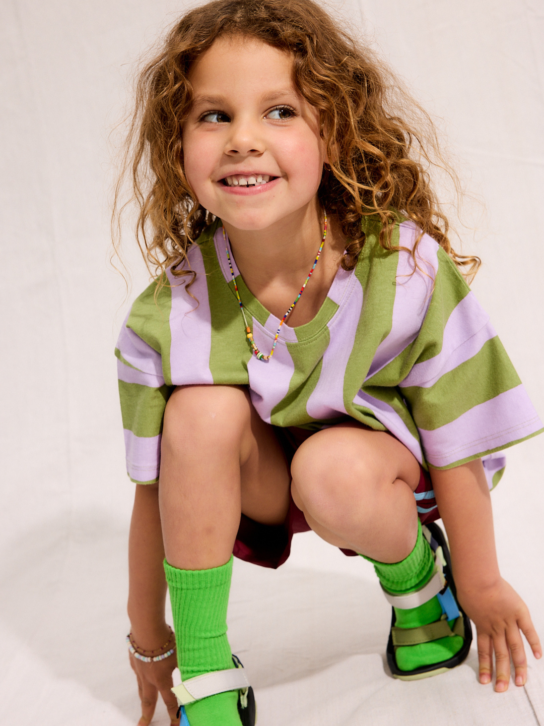 A child is wearing the Umbrella Stripe Set with green socks and sandals while squatting and smiling