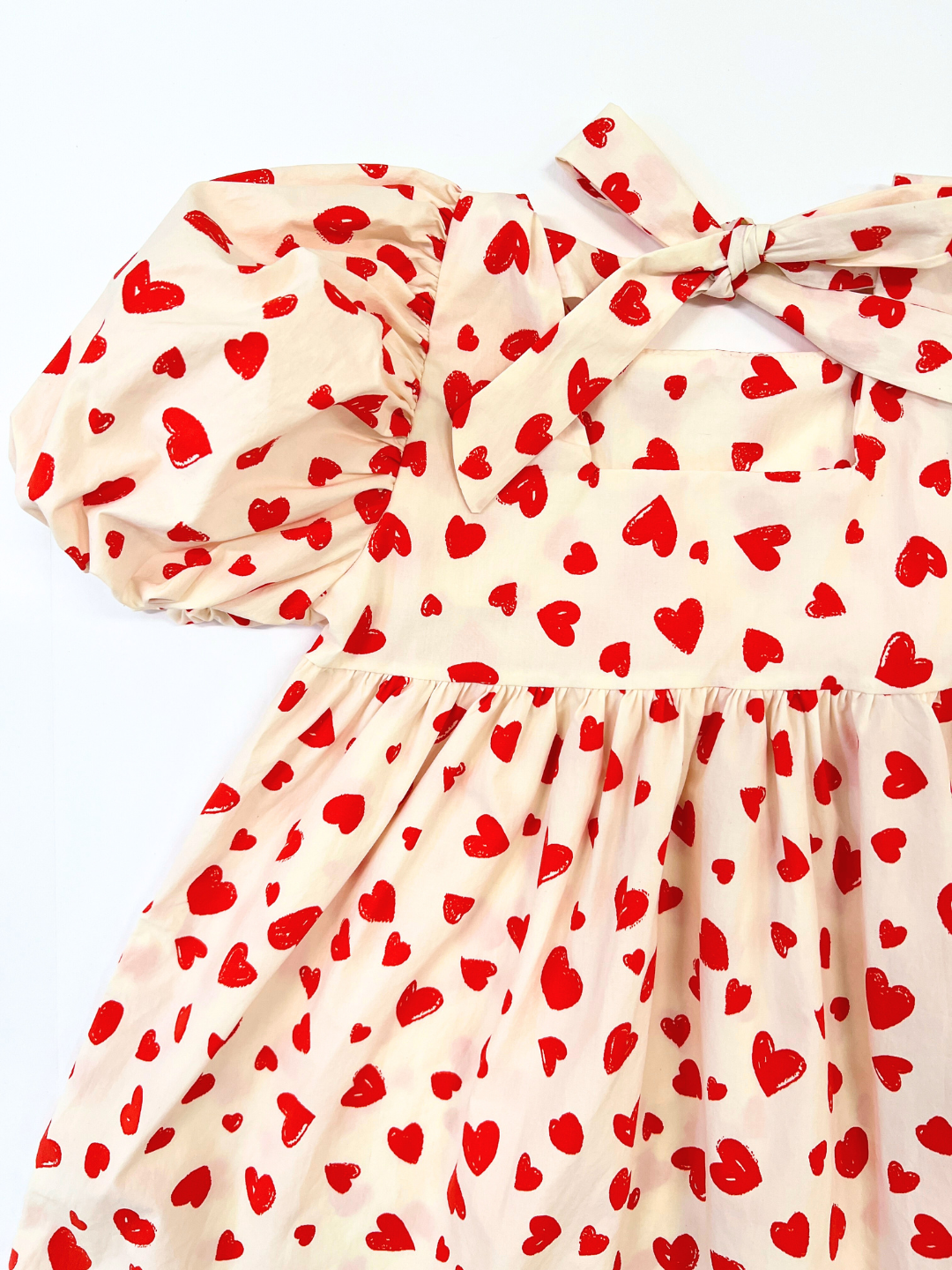 Close-up of a kids' puff-sleeved dress with red hearts on a white background, showing bow tied at the neck, back view