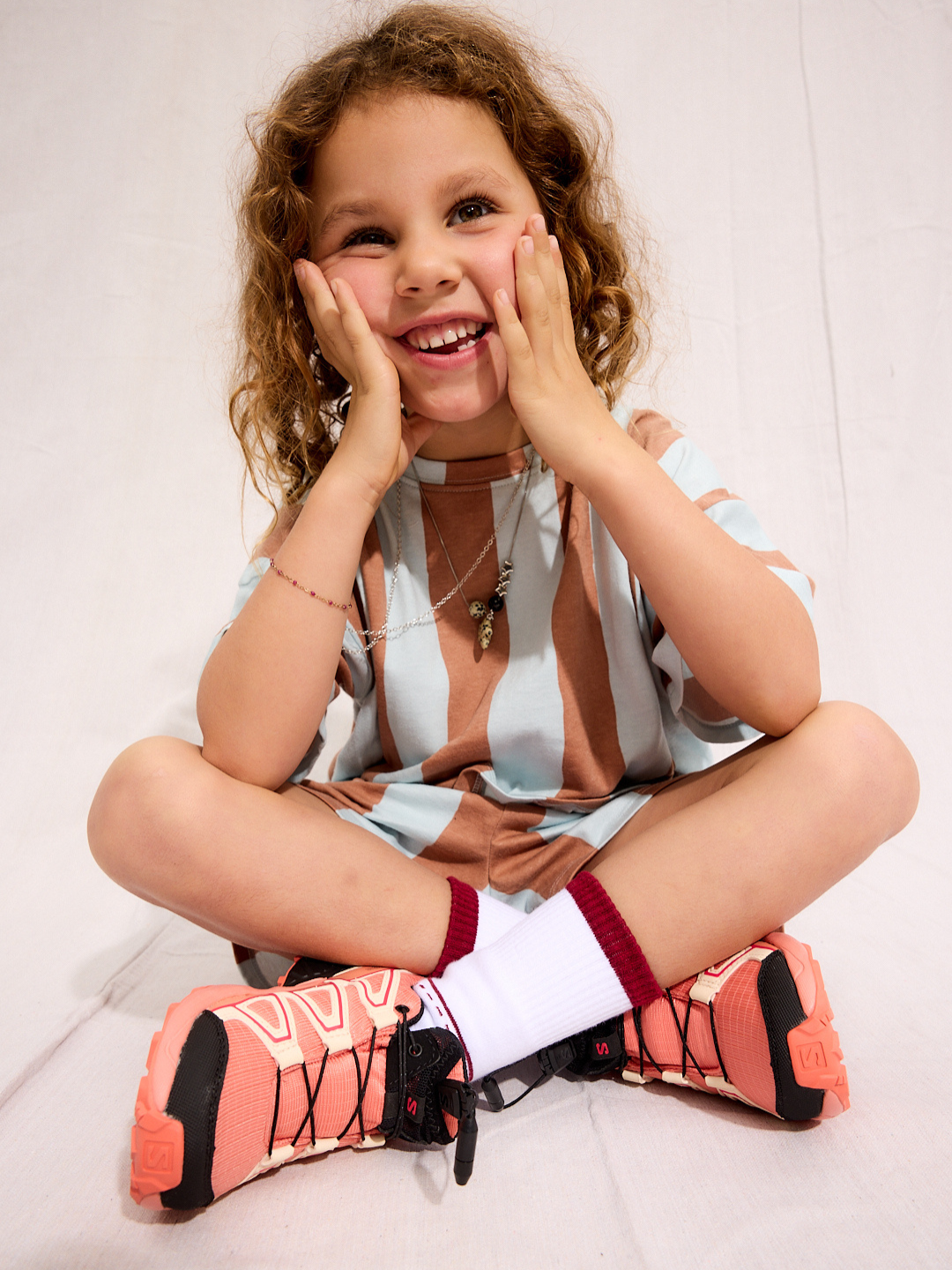 A child is wearing un Umbrella Stripe Set with white socks and red sneakers seating down and smiling