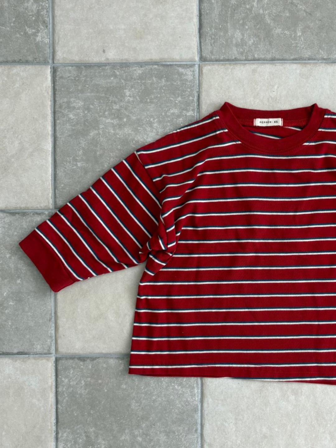 Red | The PEPPER LONGSLEEVE TEE, a red cotton top with white and blue stripes from a Korean kids brand, is laid flat on a tiled gray and beige floor.