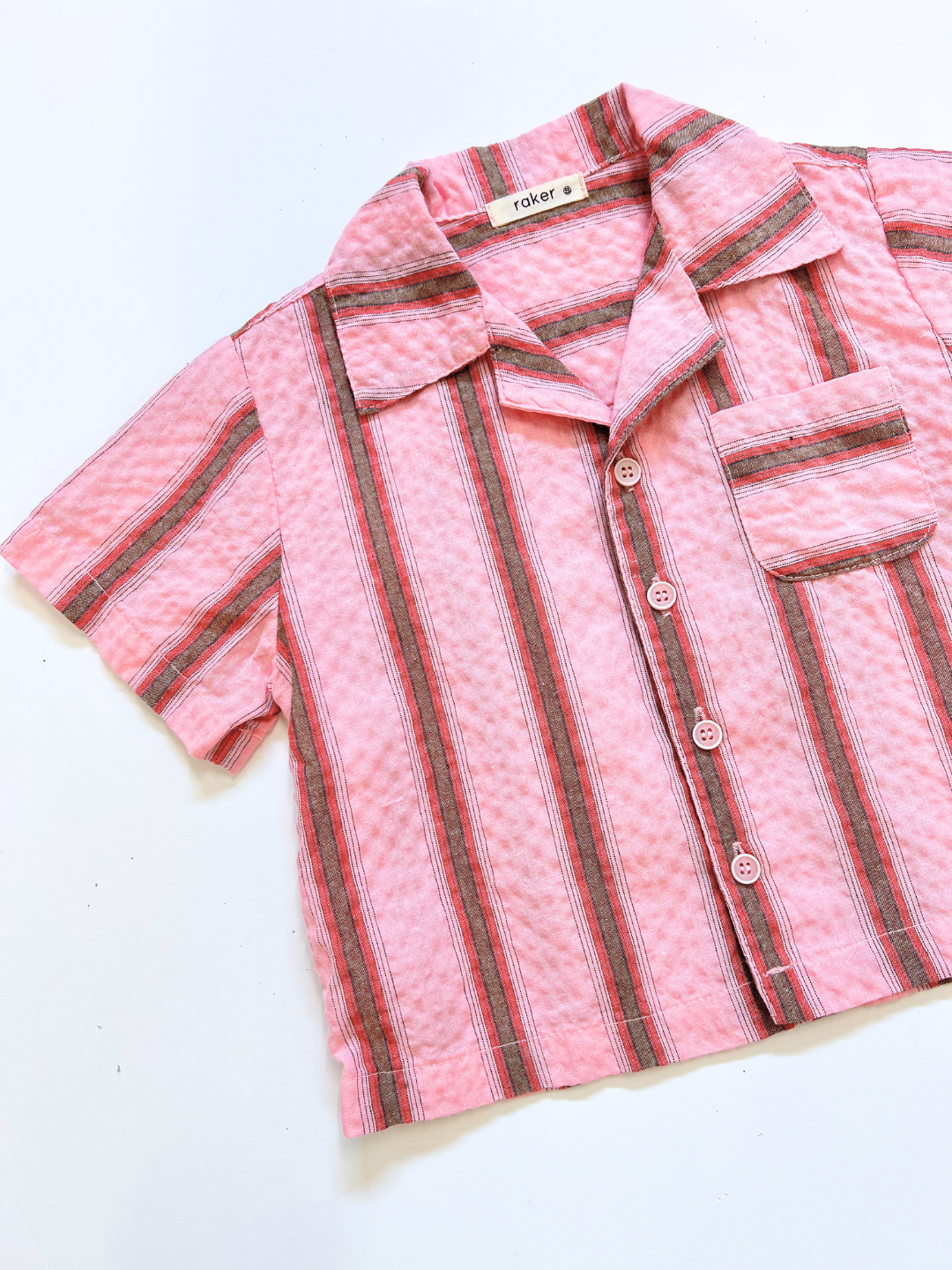 The HAVANA STRIPE SHIRT is a vintage-inspired, short-sleeve cotton shirt with pink, red, and gray vertical stripes, a chest pocket, and a classic collar.
