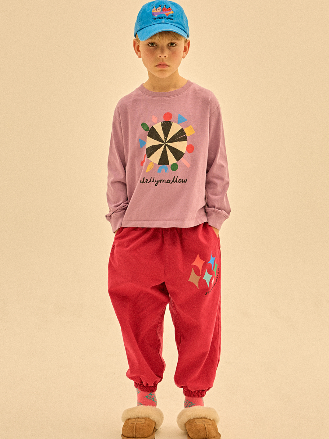A young boy stands facing forward, wearing a blue cap, the COLOR ROULETTE LONGSLEEVE T-SHIRT with a graphic design, red pants with colorful shapes, and tan slippers