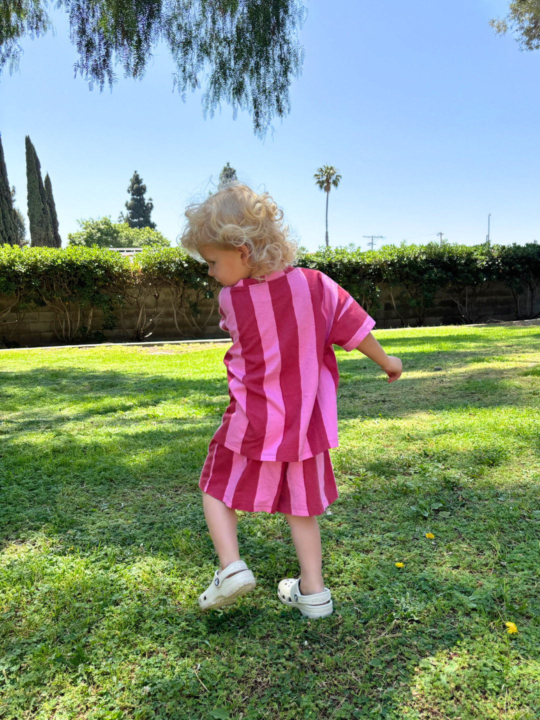 A young child walks on grass, wearing the UMBRELLA STRIPE SHORT SET in pink and red vertical stripes, made from soft organic cotton, paired with beige crocs, and blue sky behind.
