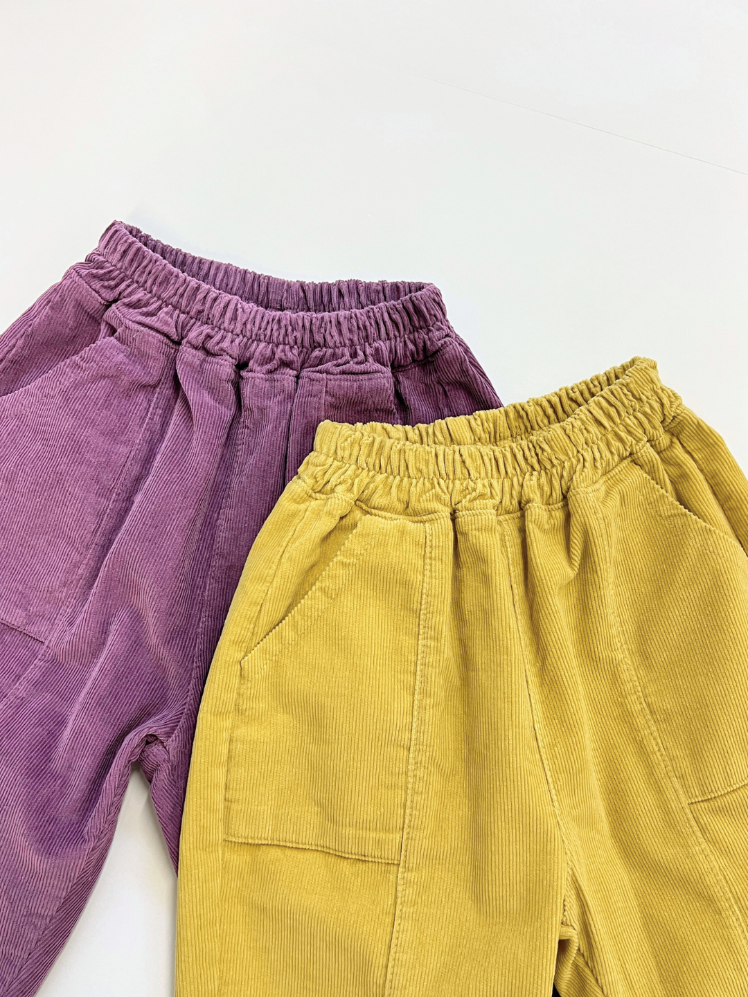 Two pairs of CORDUROY PANEL PANT from a Korean kids brand—one purple, one yellow—are laid flat side by side on a white surface.