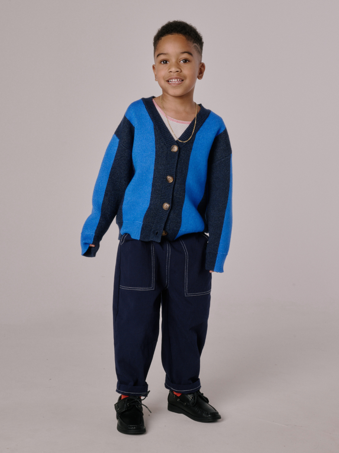 Blue | A young boy smiles while wearing the STRIPE CARDIGAN in blue and navy, navy pants with white stitching, and black shoes, standing against a plain light background.