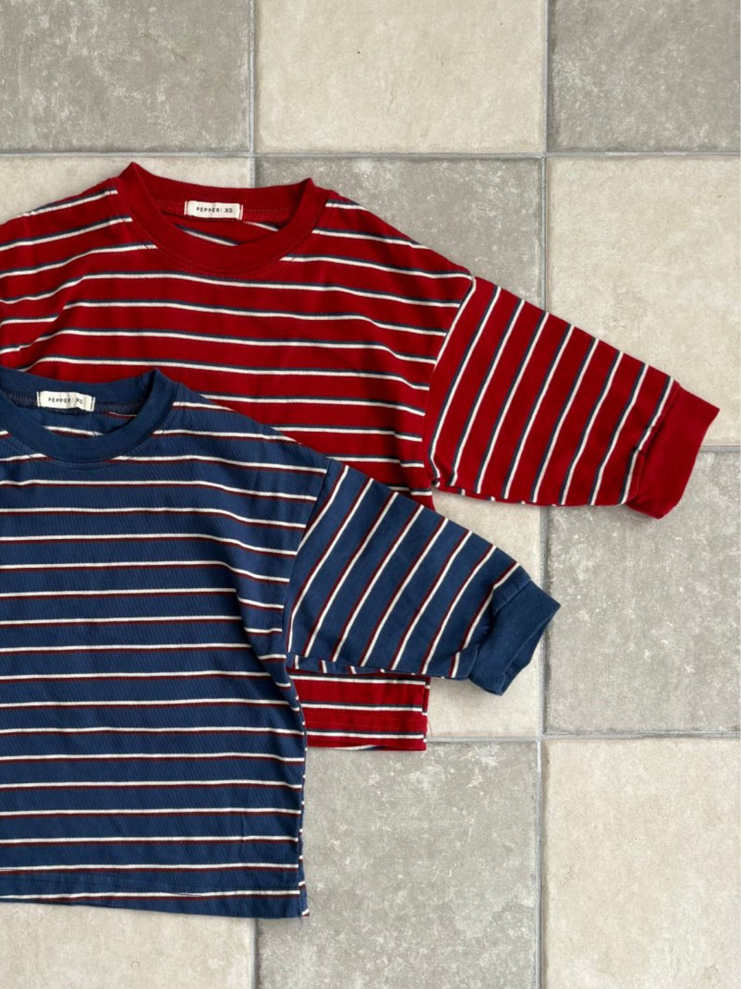 Red | Two PEPPER LONGSLEEVE TEE shirts—one red with white stripes, one navy with white stripes—from a popular Korean kids brand are neatly laid out on a tiled floor.