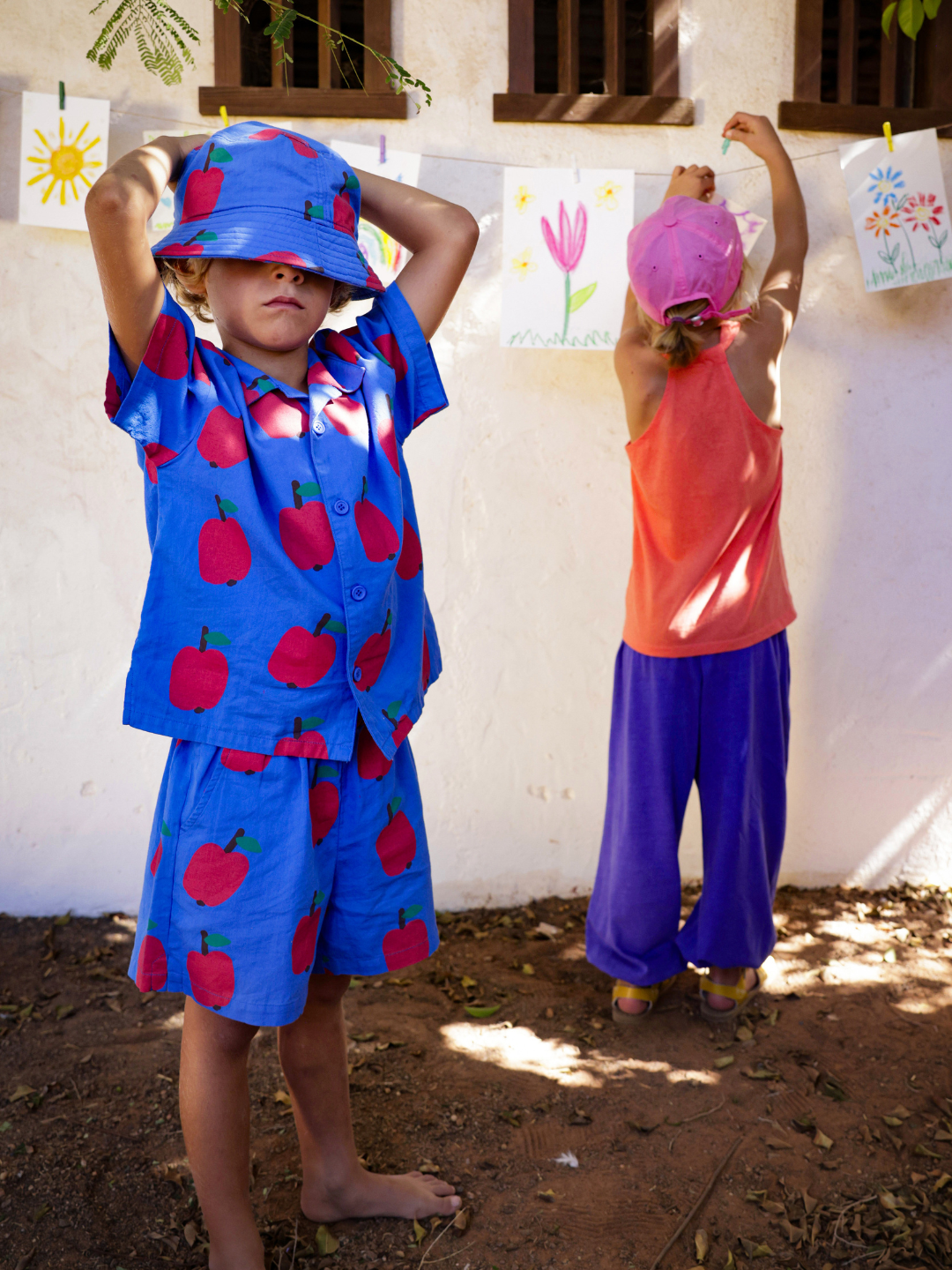 Two children hang drawings outside. One child, wearing APPLE SHORTS, covers their eyes with a hat, while the other, in a pink cap, reaches up to pin artwork.