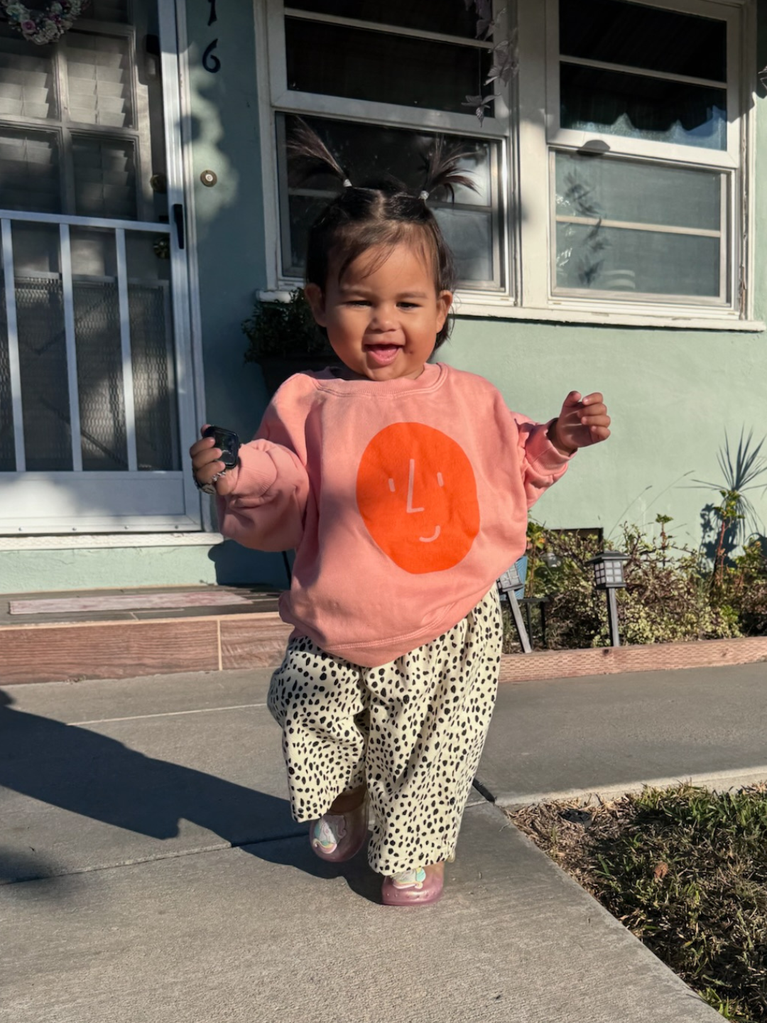 Child wearing a pink sweatshirt with an orange circle and white polka dots in front of a house.