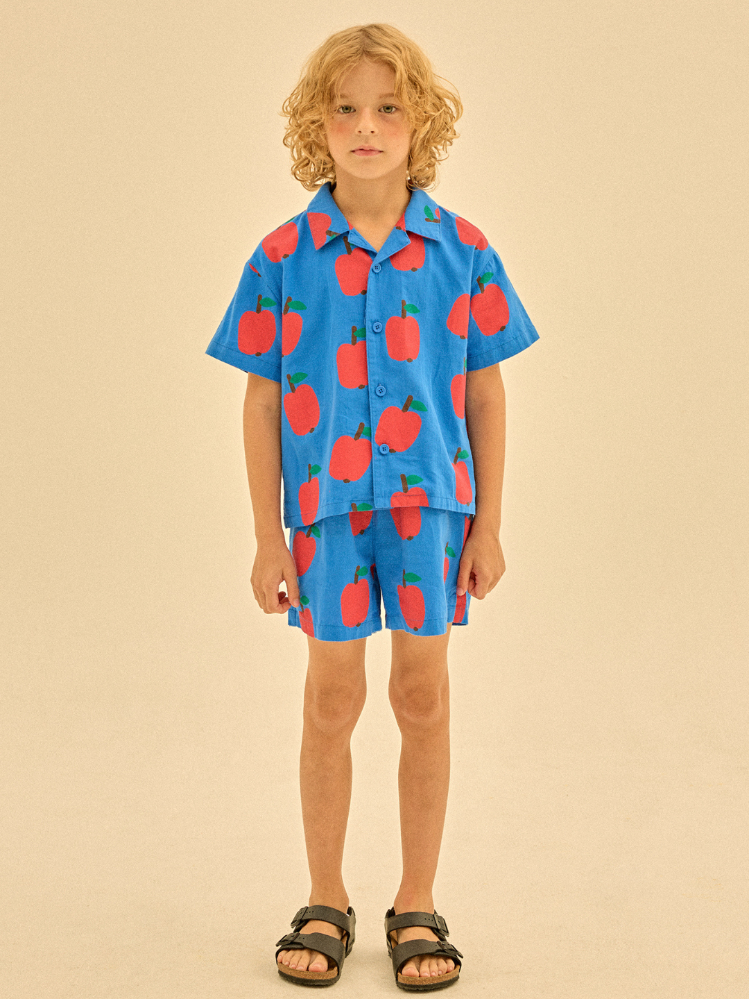 A child in the Apple Shorts set, featuring a blue shirt and shorts with red apple prints, stands against a plain beige background.