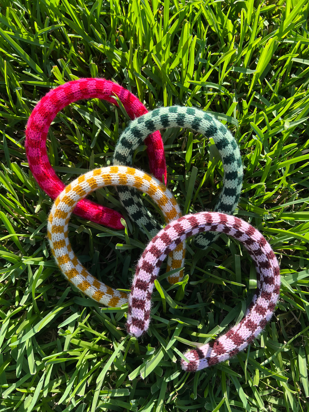 Four PUFFY KNIT HEADBANDS with checkered patterns in pink, green, yellow, and purple are arranged on green grass. Each headband is circular and has a distinct color combination, creating a vibrant and colorful display against the natural background. They capture the charm of handmade knitted accessories perfectly.