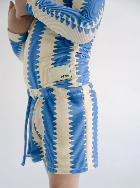 Second image of SWIM SHORTS featuring a blue and white zigzag pattern, elastic waistband with front tie, and a small rectangular label on the left leg, made from recycled material.
