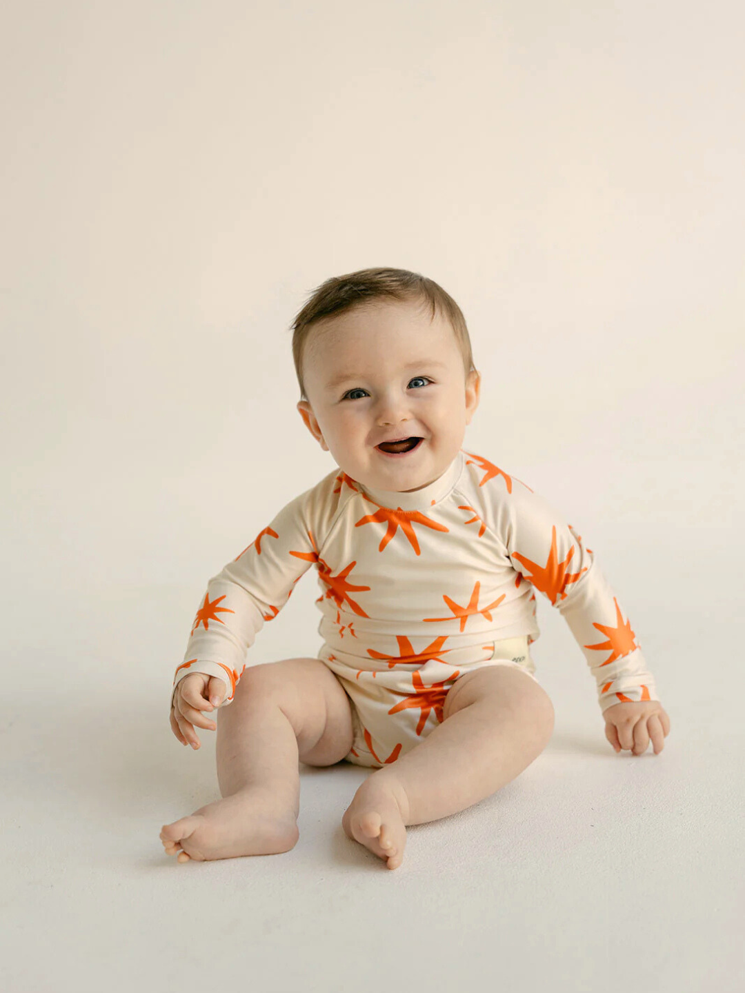 Capri | A baby sits smiling in a SWIM RASHIE, a long-sleeve onesie featuring an orange star pattern, made from soft, Oeko-Tex certified materials against a neutral background.