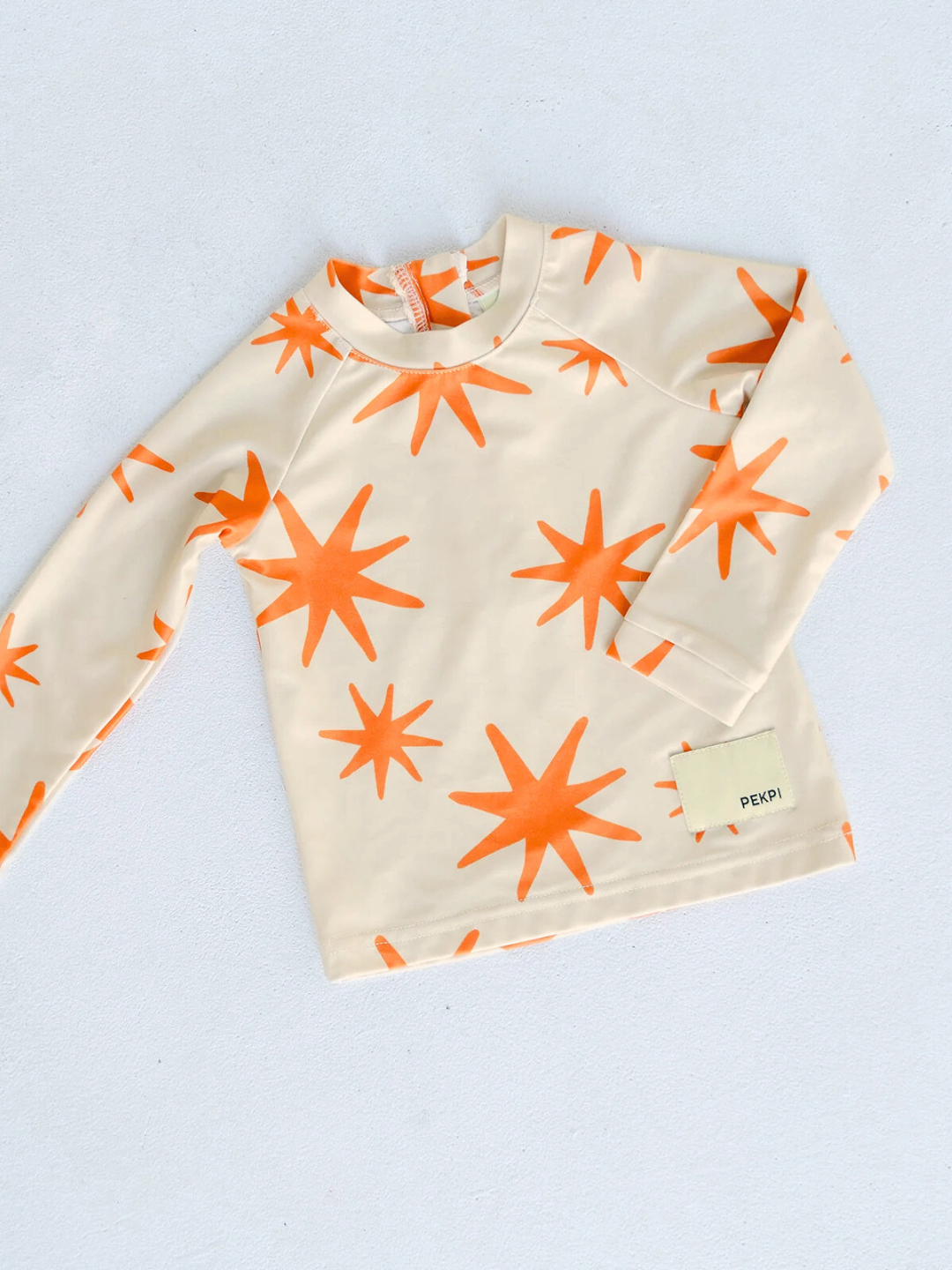 Capri | The SWIM RASHIE, a long-sleeve shirt made from recycled fabric, features an orange star design on a beige background and is displayed flat on a light surface.