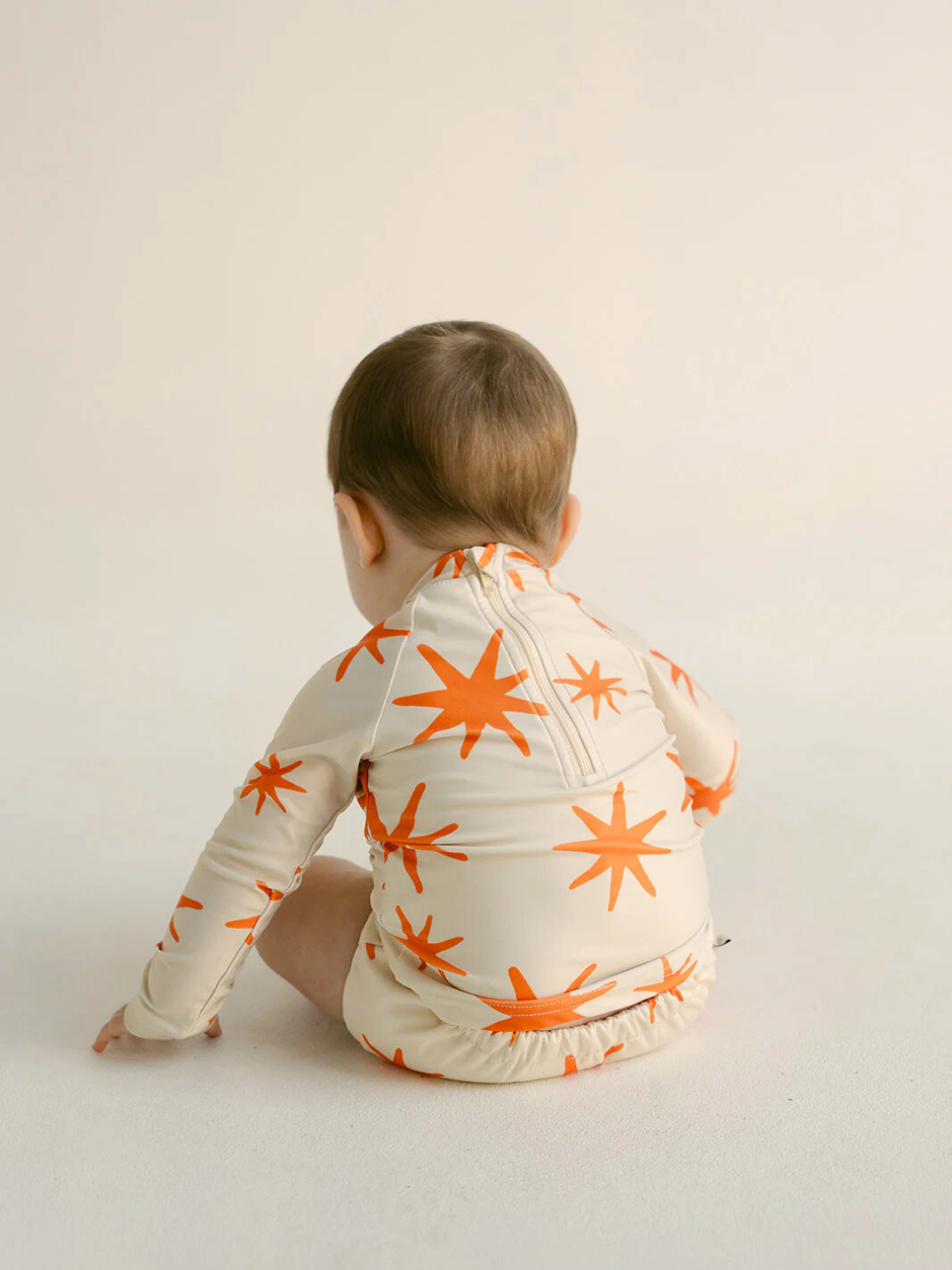Capri | A baby sits on a plain surface, facing away, wearing the SWIM RASHIE, a long-sleeve swimsuit made from recycled fabric with an orange star pattern.