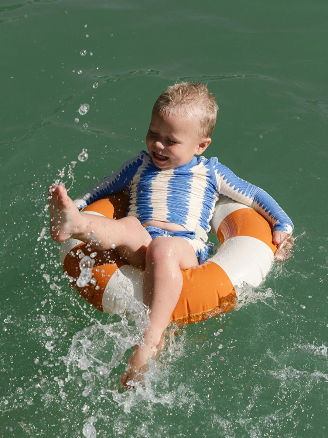 A young child in a blue and white KIDS RASHIE TOP sits in an orange and white swim ring, splashing water with their foot in a greenish pool or lake.
