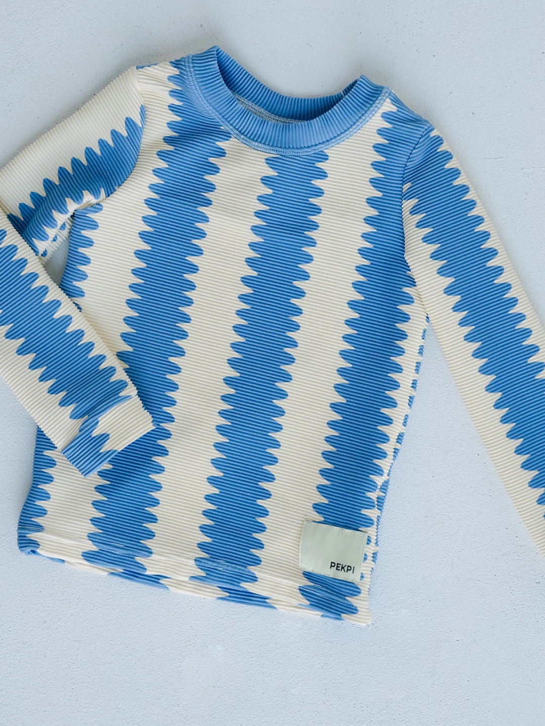 The KIDS RASHIE TOP is a long-sleeve cream shirt with blue wavy vertical stripes, a small "PEKPI" tag on the lower right, and crafted from ribbed recycled material for comfort and style, shown laid flat on a light surface.