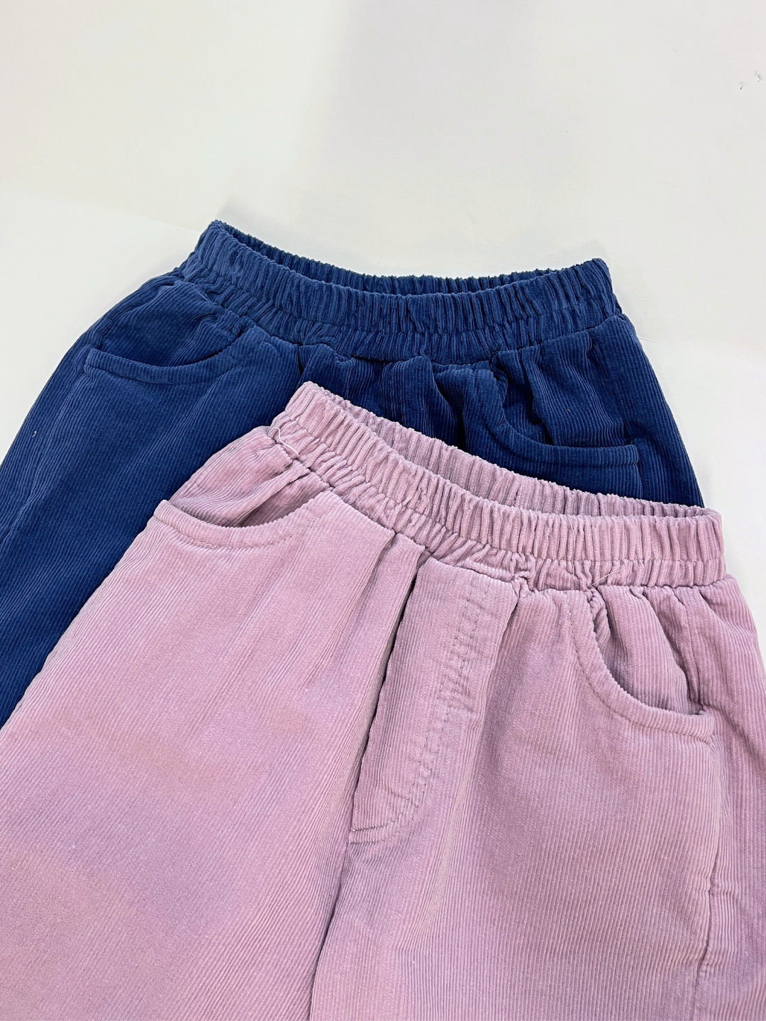 Two pairs of SIMPLE CORDUROY PANTS from a Korean kids brand—navy blue and light pink—made of cotton corduroy with elastic waists and side pockets, are displayed on a white background.