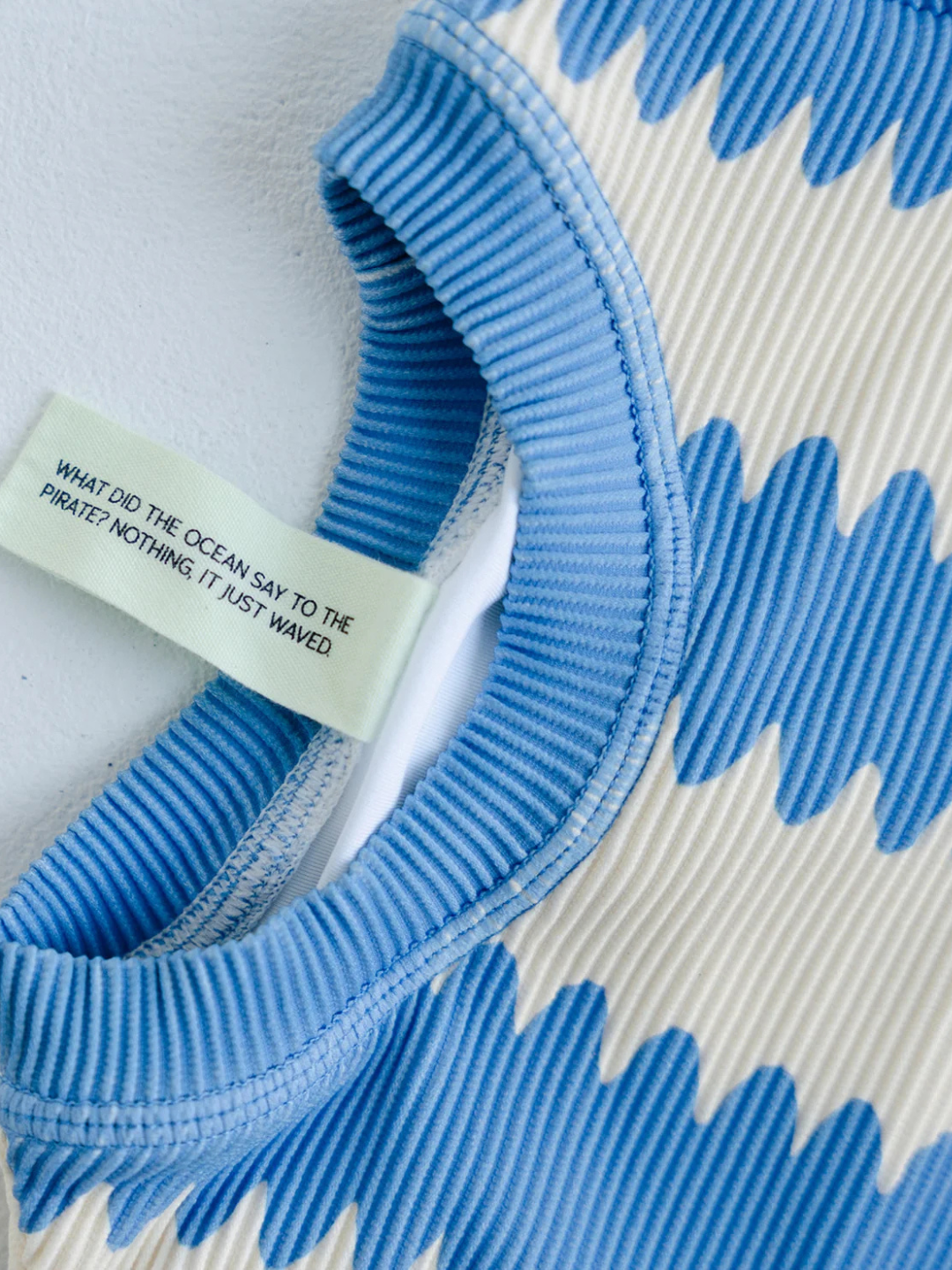 Close-up of the KIDS RASHIE TOP, a blue and white ribbed long-sleeve made from recycled material. Label reads: "WHAT DID THE OCEAN SAY TO THE PIRATE? NOTHING IT JUST WAVED." UPF 50+ for stylish sun protection.