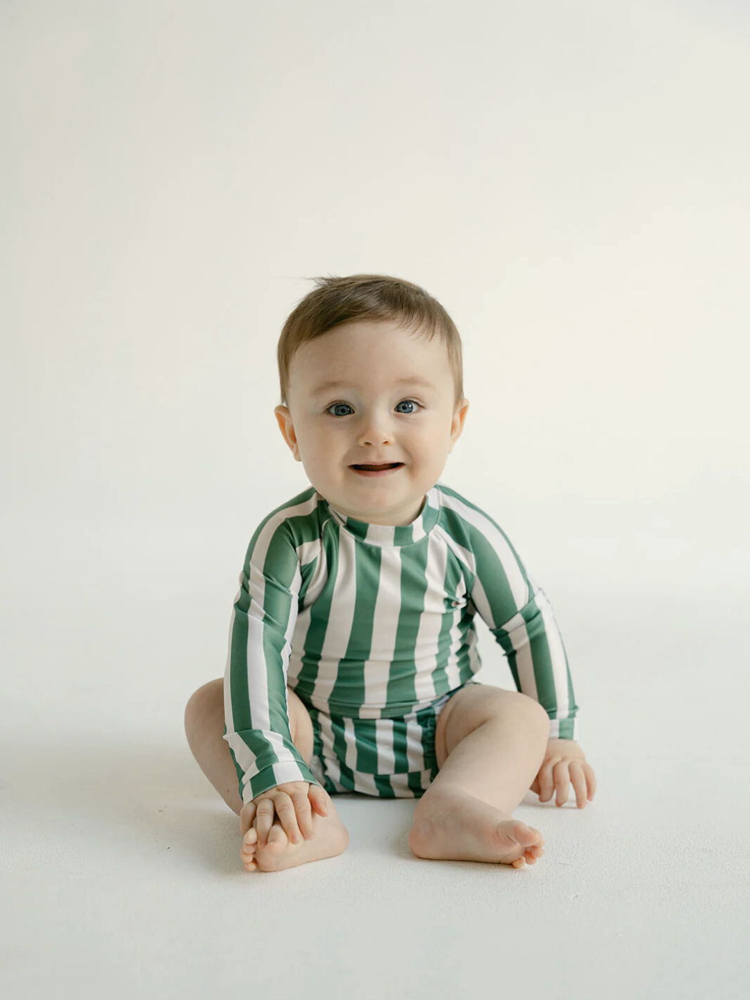 Jungle | A baby in a green and white striped SWIM RASHIE, crafted from Oeko-Tex certified fabric, sits on a white surface, gazing at the camera with a neutral expression.