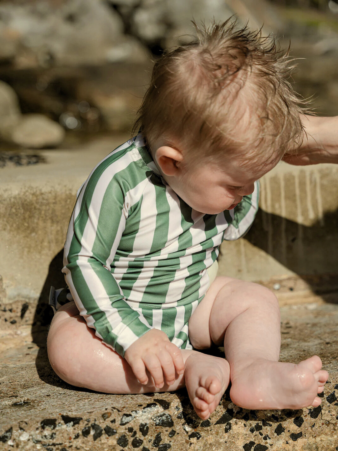 Jungle | A baby wearing the SWIM RASHIE, a green and white striped swimsuit crafted from recycled fabric, sits with wet hair against a stone surface and water backdrop, creating a serene scene.
