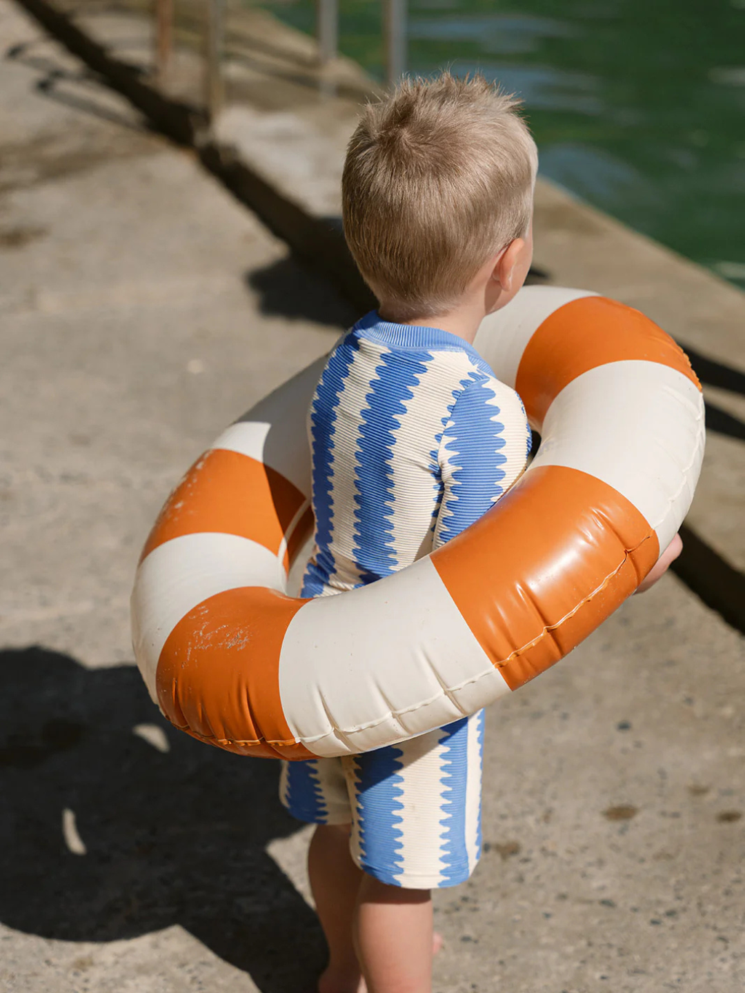 A young child wearing a blue and white striped KIDS RASHIE TOP with UPF 50+ stands on concrete, holding a large orange and white inflatable swim ring, facing the water.