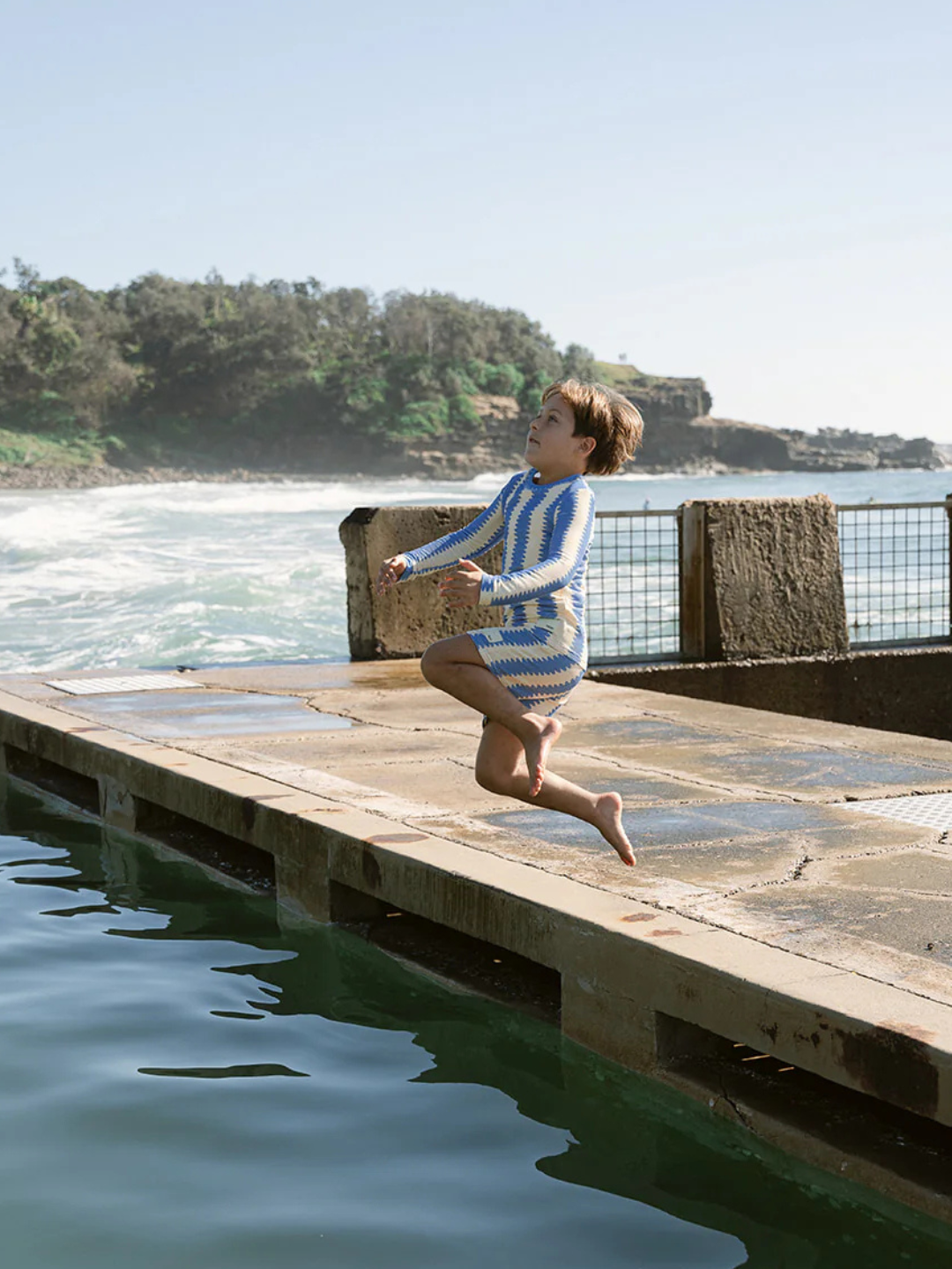 Wearing SWIM SHORTS made from recycled material, a person jumps mid-air near a concrete pier by the ocean, with waves and greenery in the background.