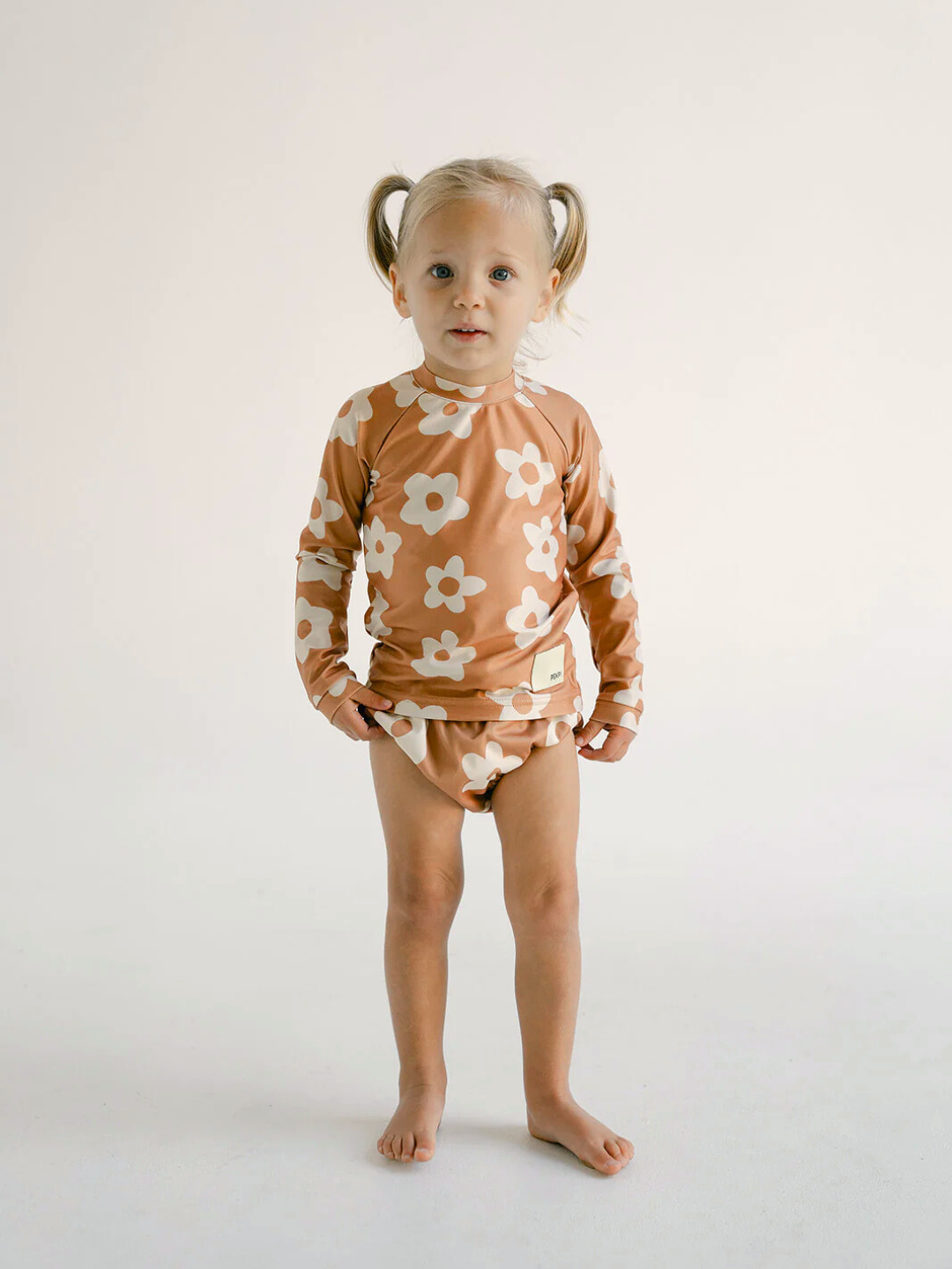Posy | A young child with pigtails stands against a plain background, wearing the charming SWIM RASHIE, a long-sleeve shirt with a floral pattern and matching diaper cover made from recycled fabric.