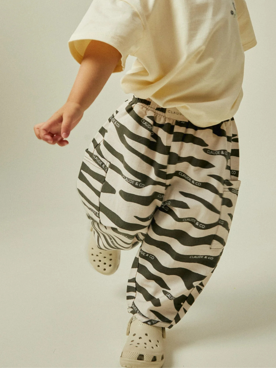 A child wearing the EMERSON ZEBRA TROUSER, featuring a wavy black and white design with text, walks in a cream-colored shirt. The baggy pants' oversized silhouette is paired with beige shoes featuring holes, enhancing the casual charm of this organic cotton outfit.