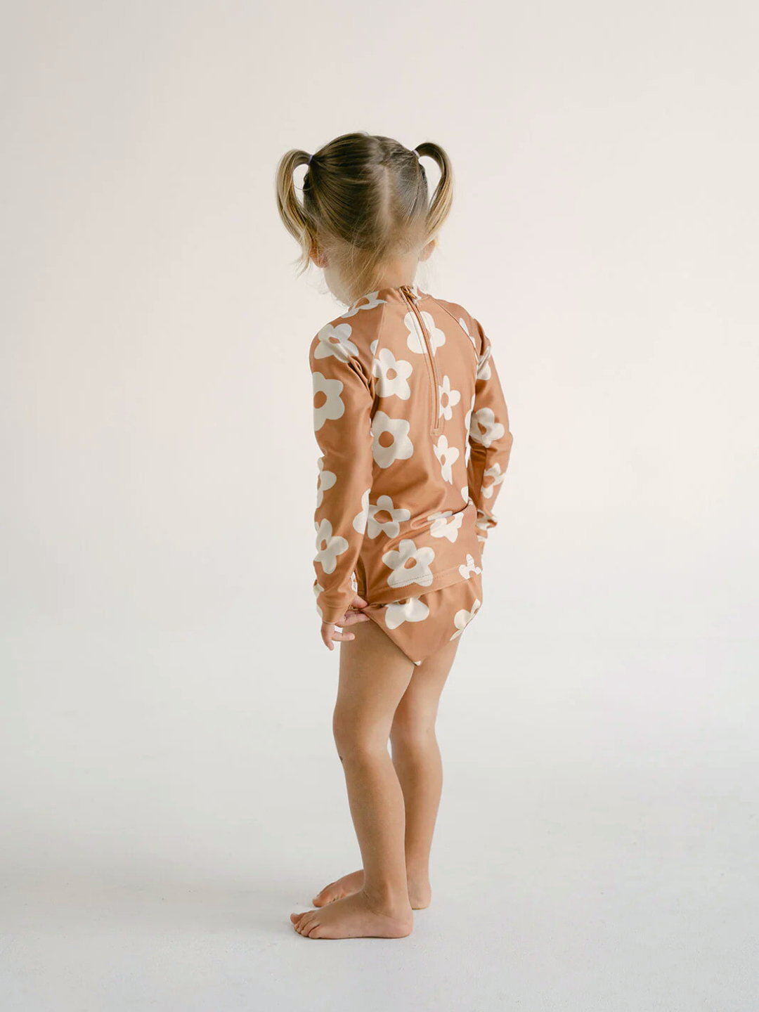 Posy | A child with pigtails, wearing the SWIM RASHIE—a floral-patterned swimsuit made from recycled fabric—stands barefoot against a plain background.