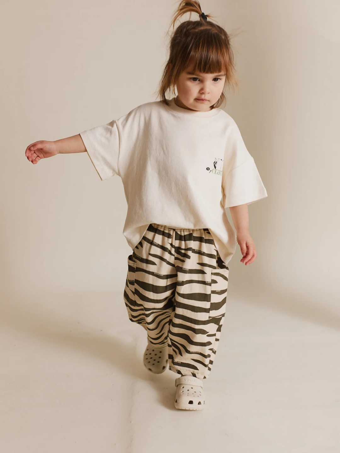 A young child strolls in an oversized silhouette, wearing a white organic cotton t-shirt, paired with EMERSON ZEBRA TROUSER and white Crocs against a plain background.