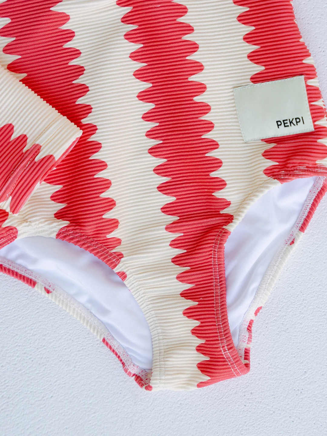 Close-up of the FULL PIECE SWIMSUIT, showcasing textured recycled fabric with red and cream zigzag stripes and a sewn-on "PEKPI" label, displayed on a light surface.
