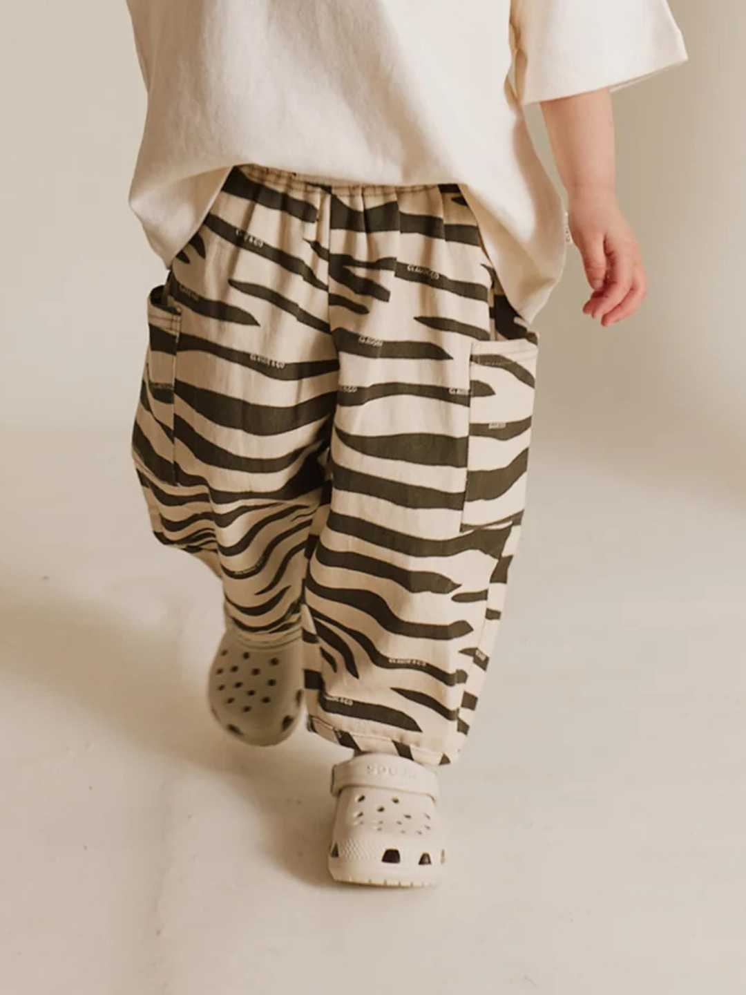 A child in a white shirt and EMERSON ZEBRA TROUSER made from organic cotton walks away on a neutral background, their white clogs softly touching the floor.