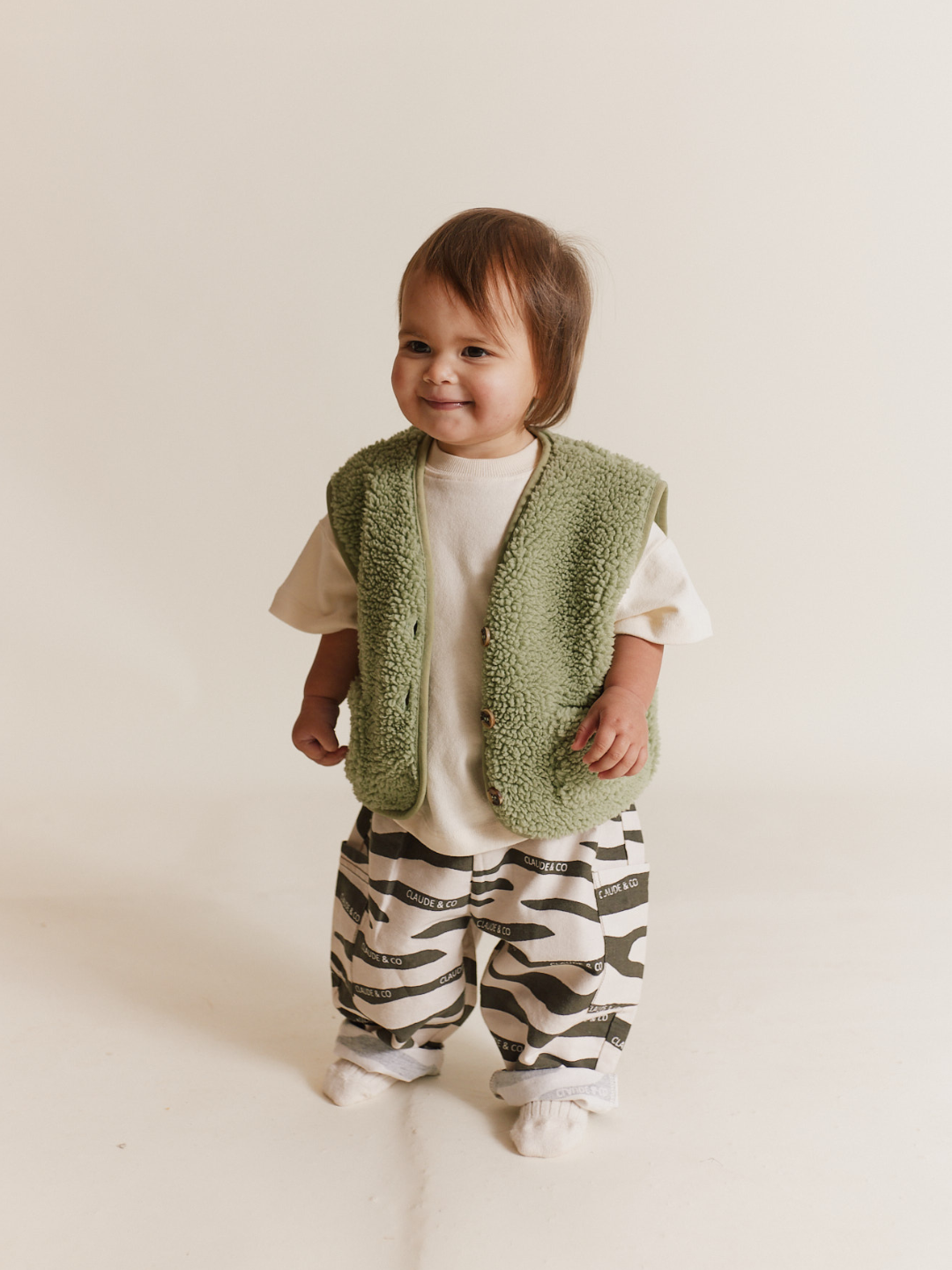 A toddler stands with a slight smile, wearing the EMERSON ZEBRA TROUSER made of organic cotton, paired with a green furry vest and a white t-shirt, against a plain background.