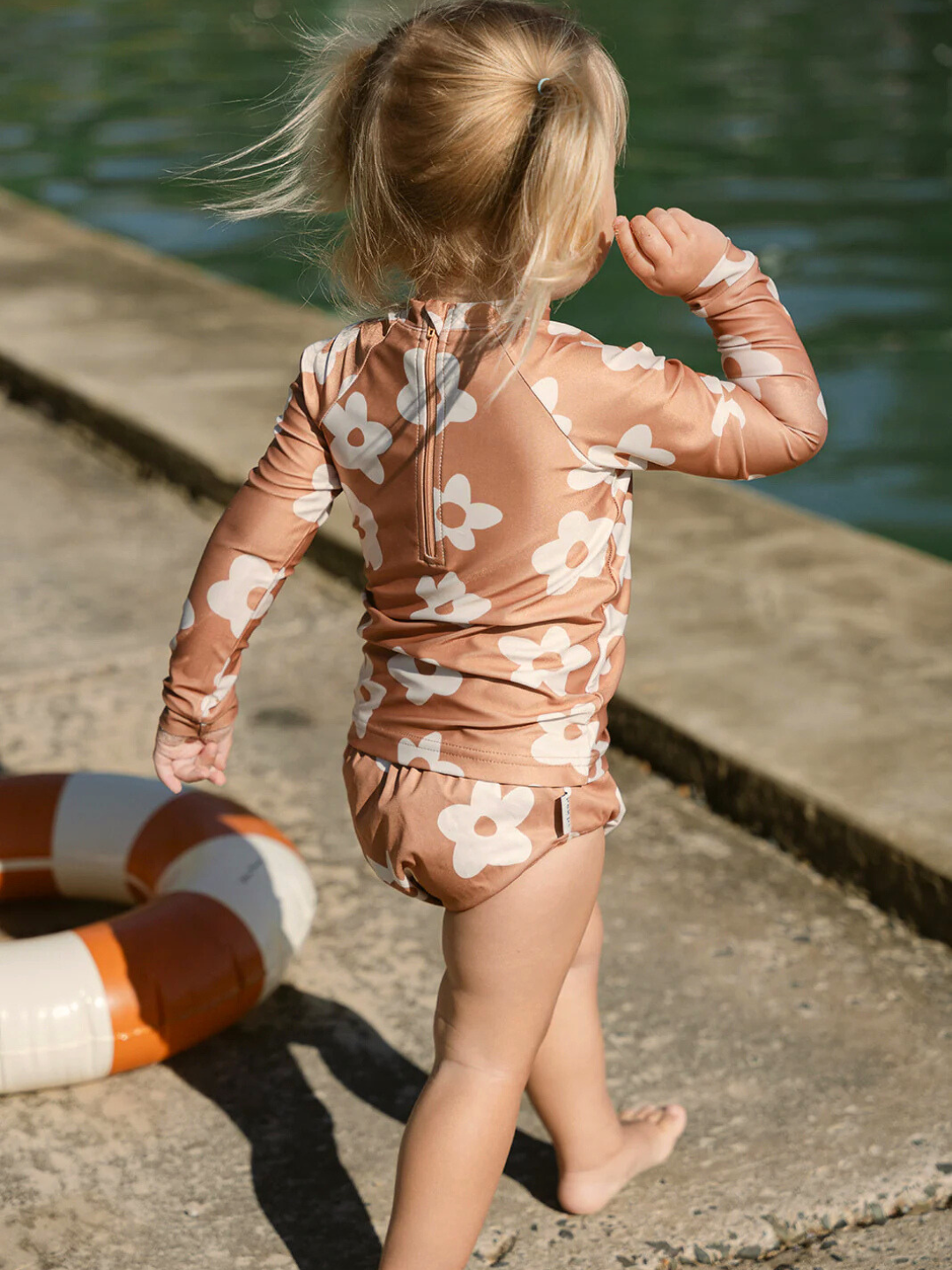 Posy | A young child stands by a concrete edge near water wearing a SWIM RASHIE, featuring a floral pattern. This swimsuit is crafted from Oeko-Tex certified recycled fabric, with a life ring on the ground nearby.