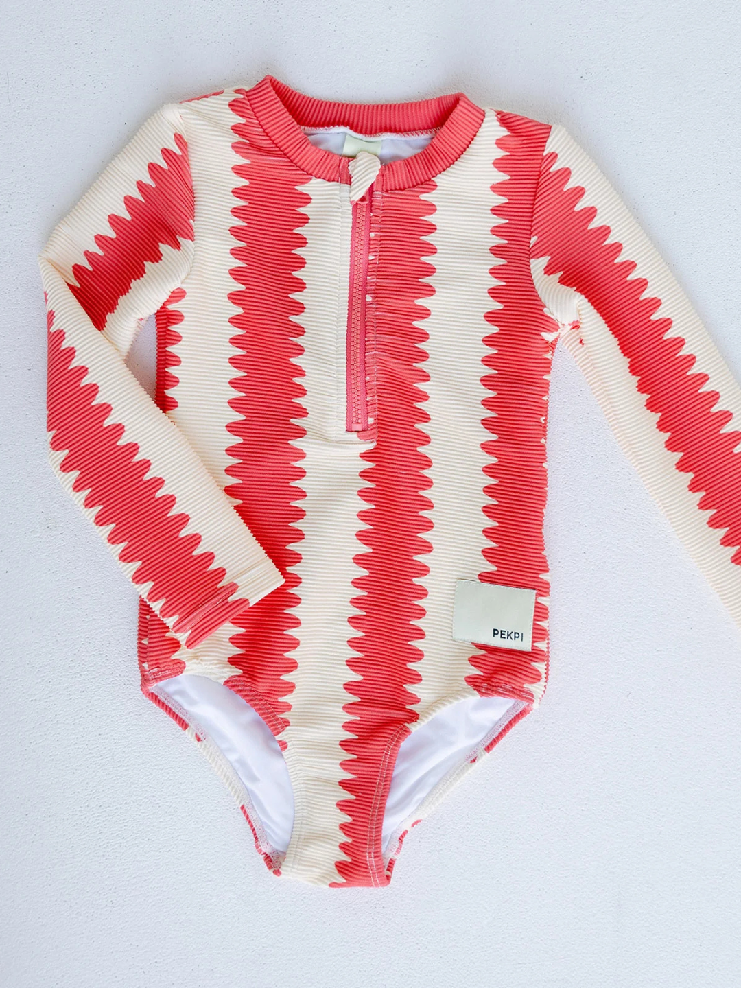 The FULL PIECE SWIMSUIT is a long-sleeve, one-piece swimsuit with a red and cream zigzag pattern, recycled fabric, front zipper, “PEEK” tag, and UPF 50+ UV protection, shown laid flat on a white surface.